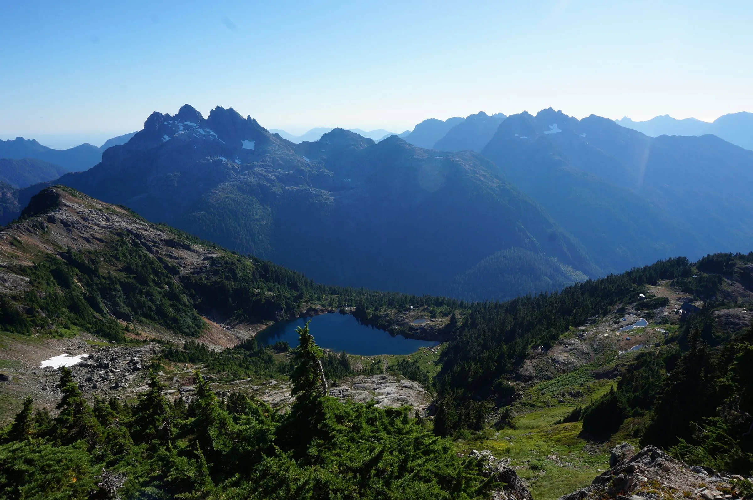 A deep blue tarn in a forested bowl below a jagged, snow-flecked peak, more ridges fading into haze