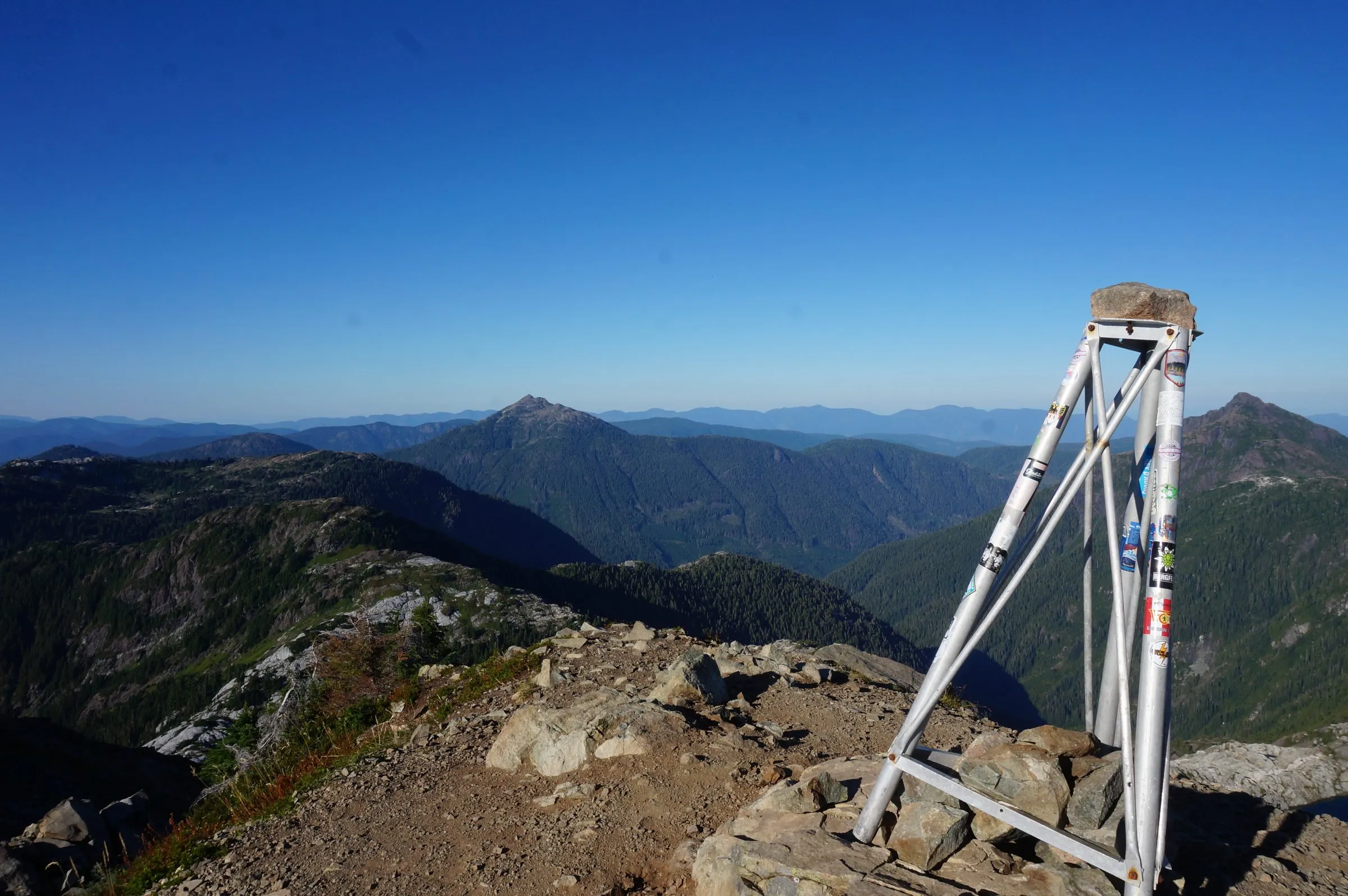A sticker-covered metal survey tripod set in a rock cairn on the summit, a pointed peak and forested valleys beyond