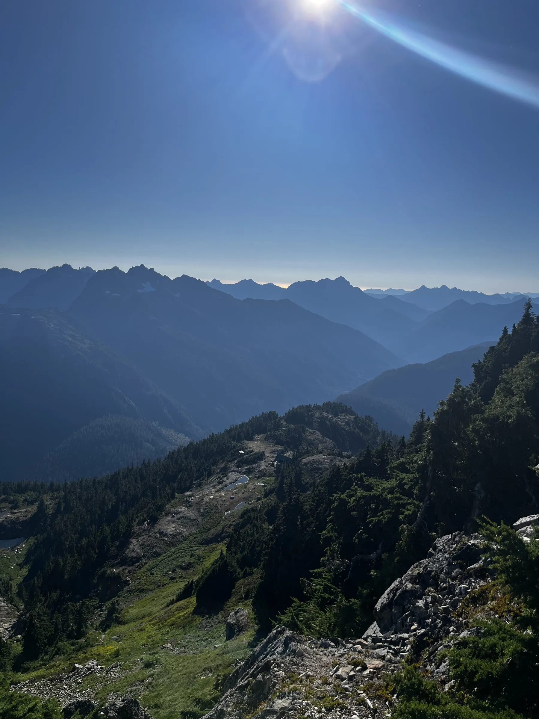 Hazy blue ridges receding under a bright sun with lens flare, a green alpine slope with small ponds below a rocky outcrop