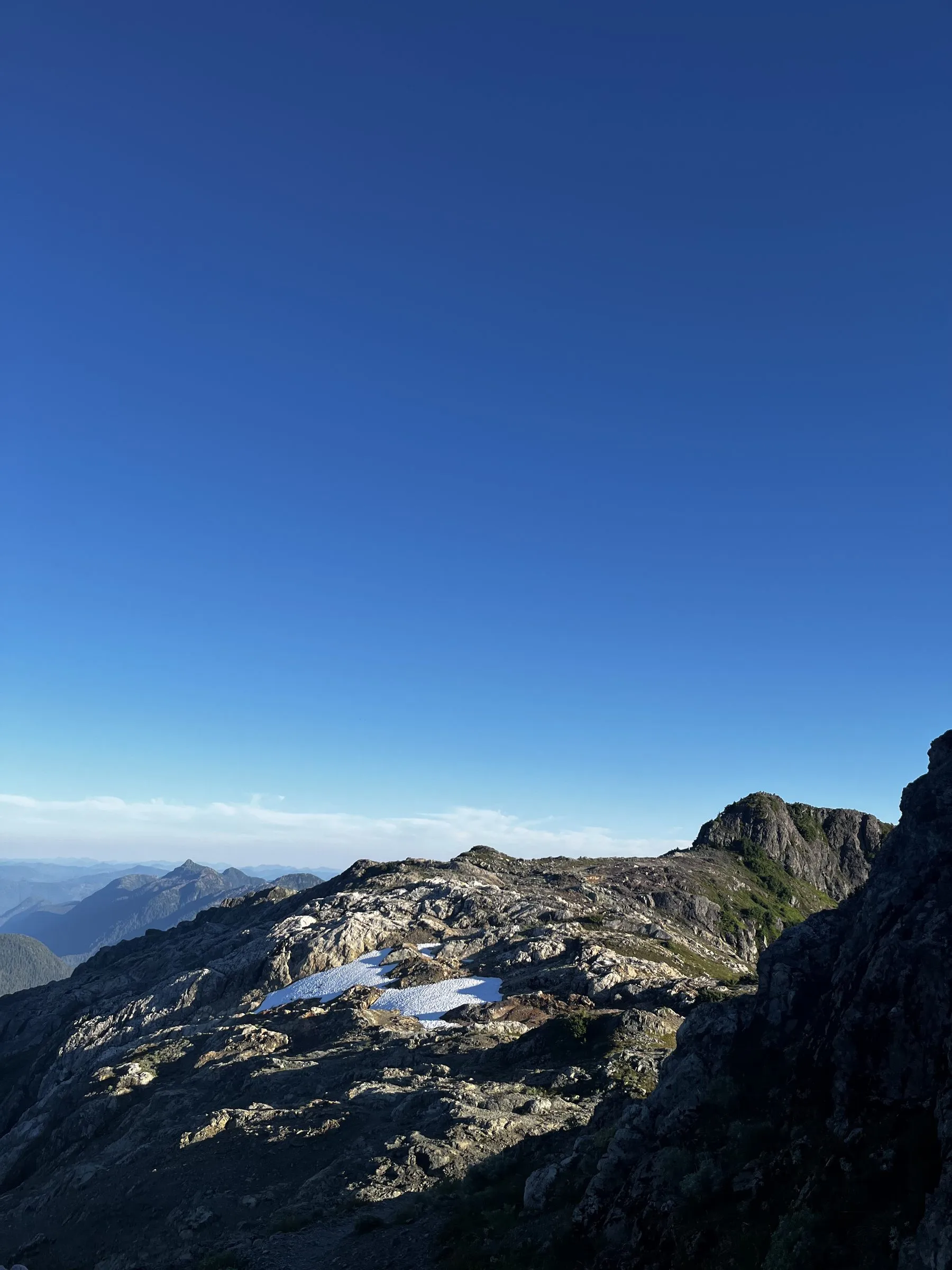 A lingering patch of snow on a rocky ridge beneath a vast, deep blue sky, distant peaks to the left