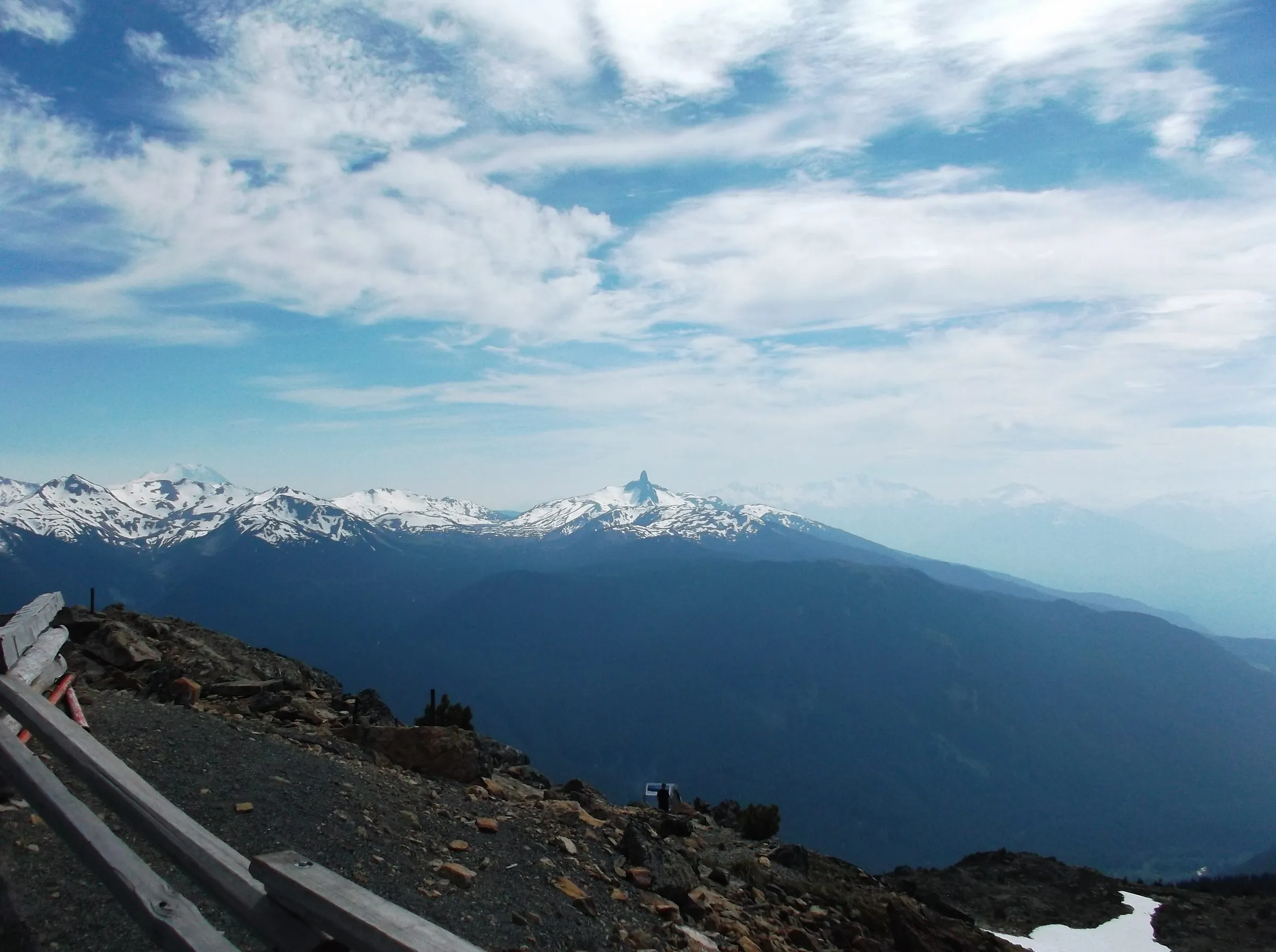 Snow-capped peaks including the spire of Black Tusk across a hazy valley, seen from a rocky summit with a wooden railing in the foreground.
