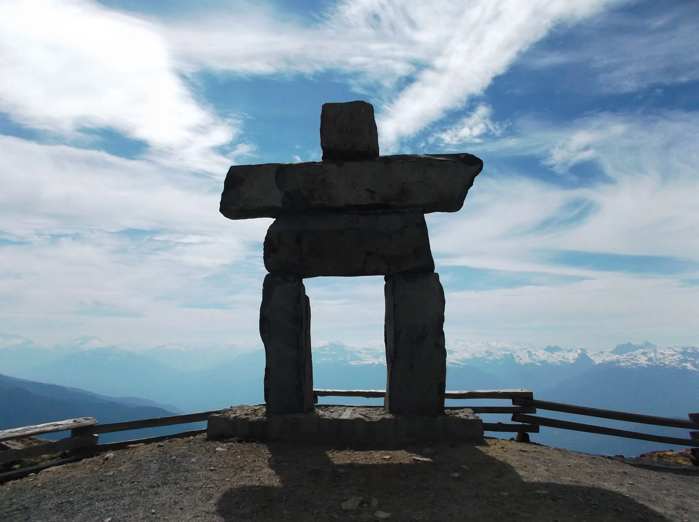 A large stone inukshuk silhouetted against a streaky sky on a mountaintop, snowy ranges along the horizon behind a wooden fence.