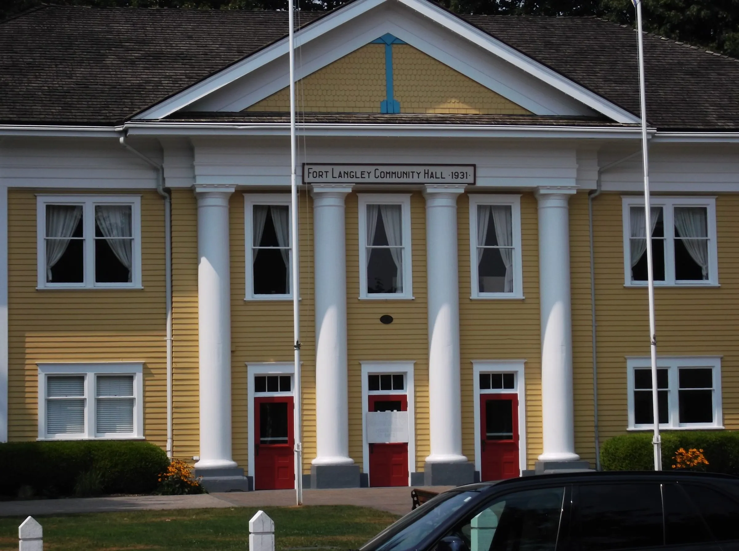 The yellow clapboard Fort Langley Community Hall with white columns, red doors and a 1931 nameplate, a dark car parked in front.