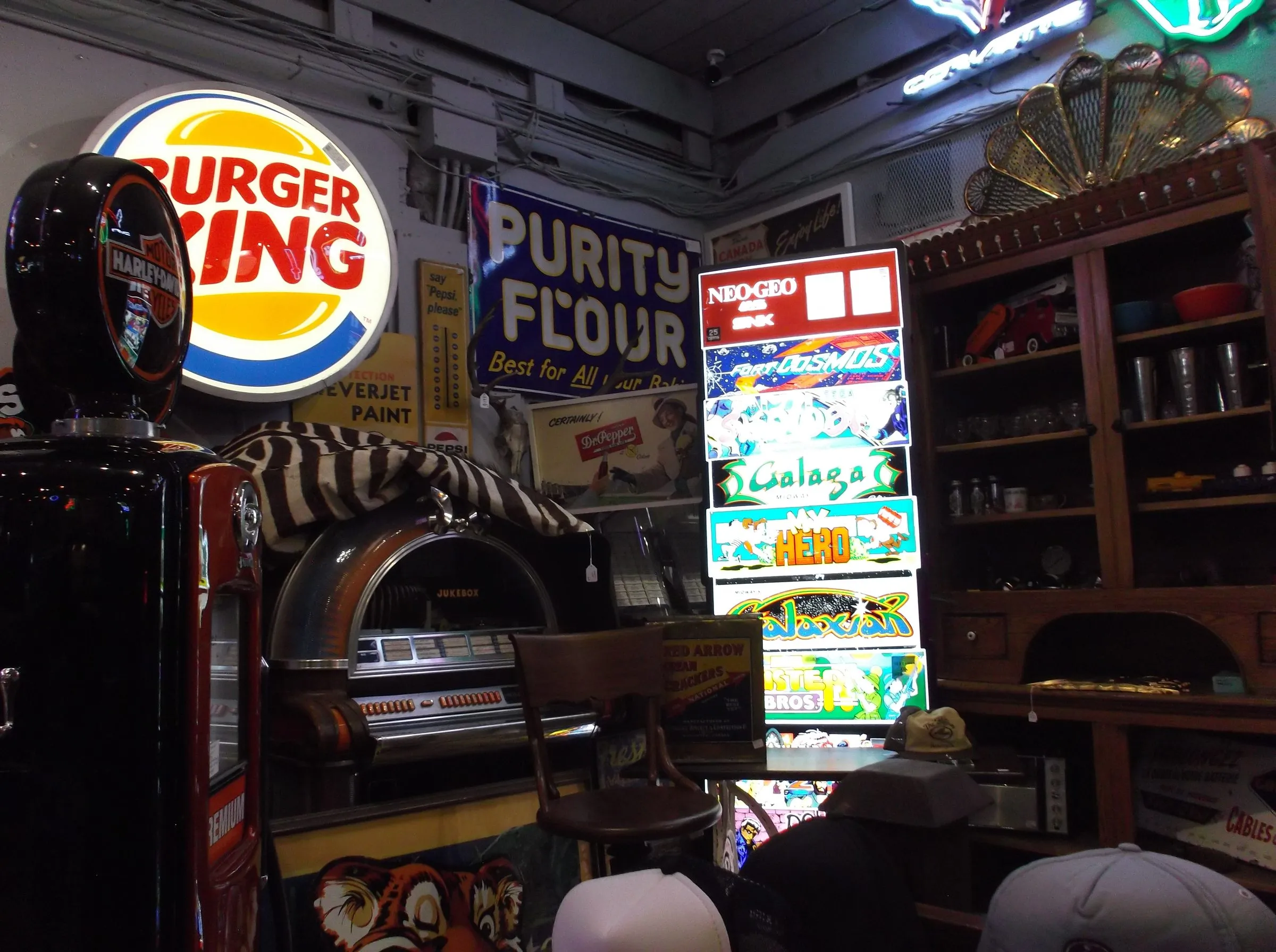 A cluttered antique shop corner with a lit Burger King sign, a jukebox, a vintage gas pump and a stack of glowing arcade marquees.