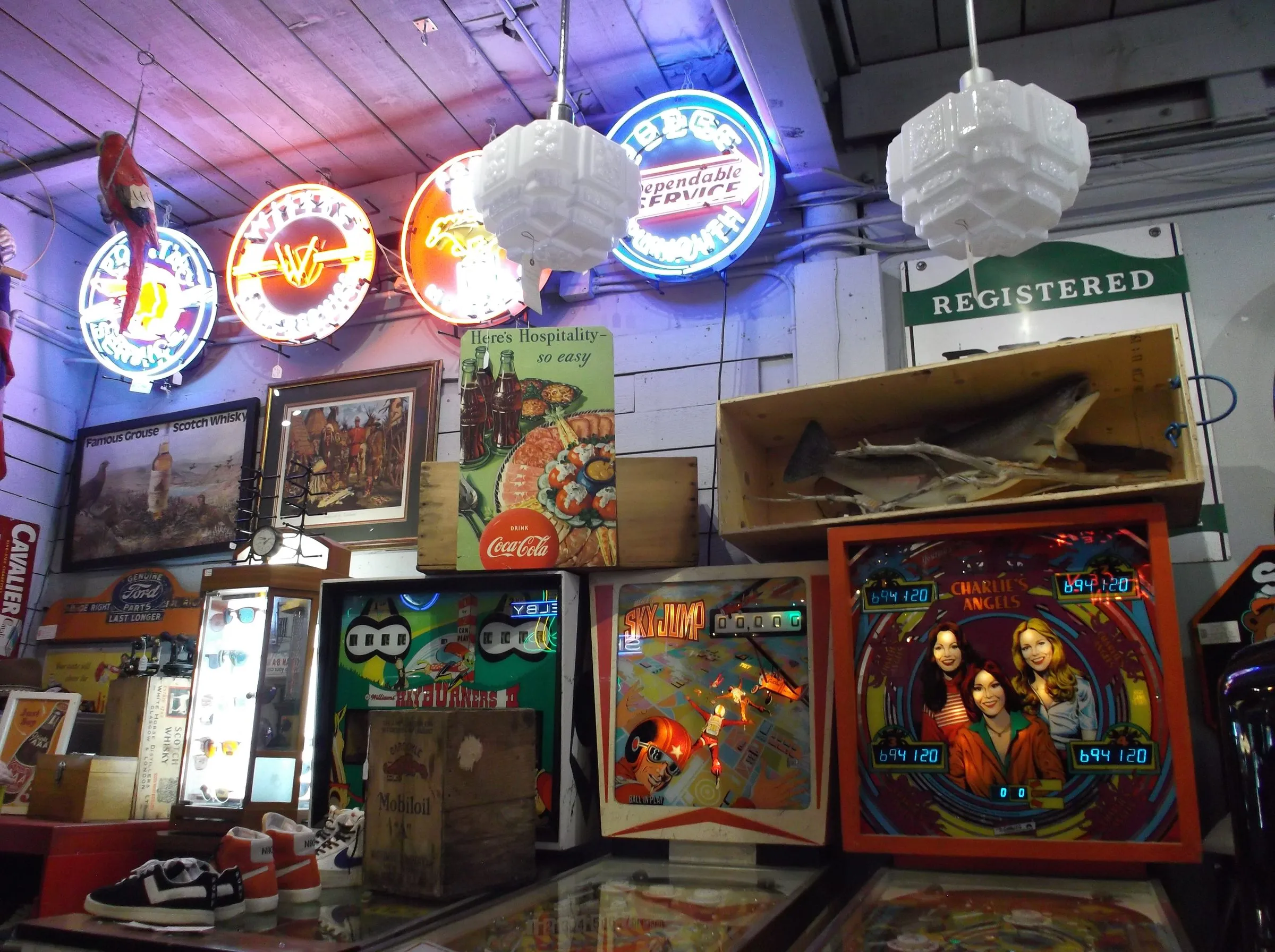 Vintage neon signs and milk-glass pendant lamps above old pinball backglasses, a Coca-Cola ad and a mounted fish in a wooden crate.