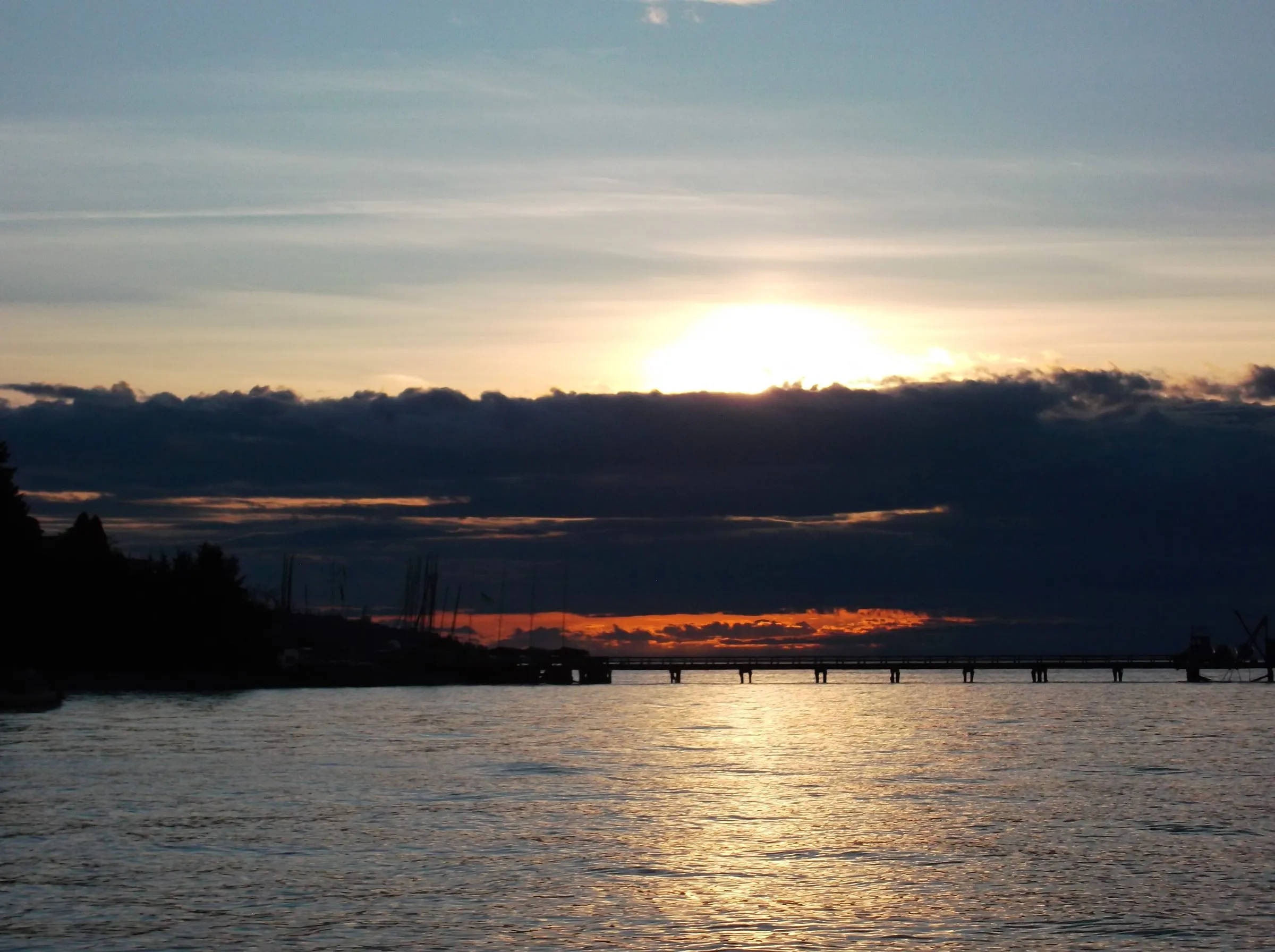 The sun setting behind a band of dark cloud over open water, a long low pier on the horizon