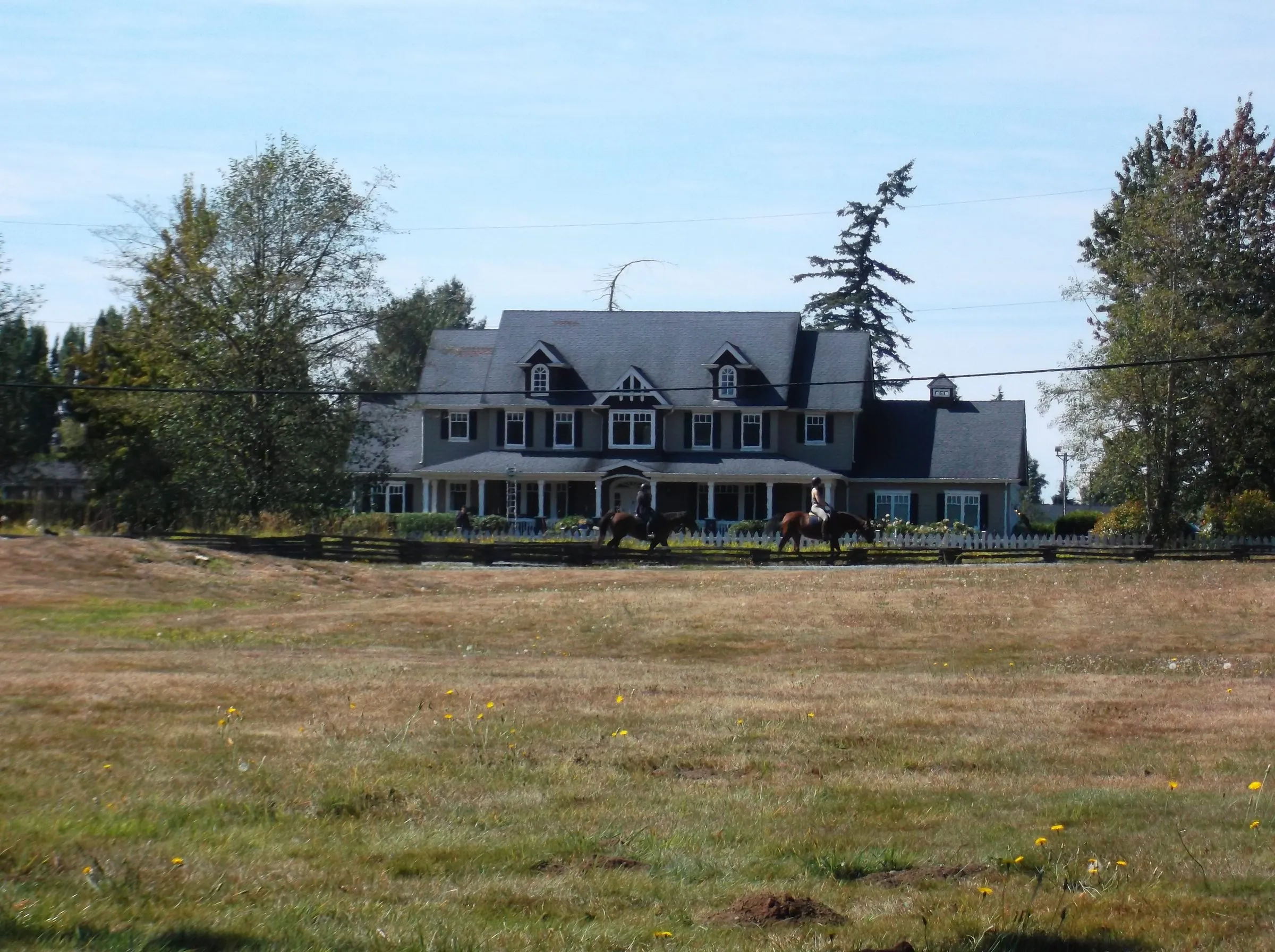 Two horseback riders pass in front of a large grey farmhouse with dormer windows, beyond a dry, open field.
