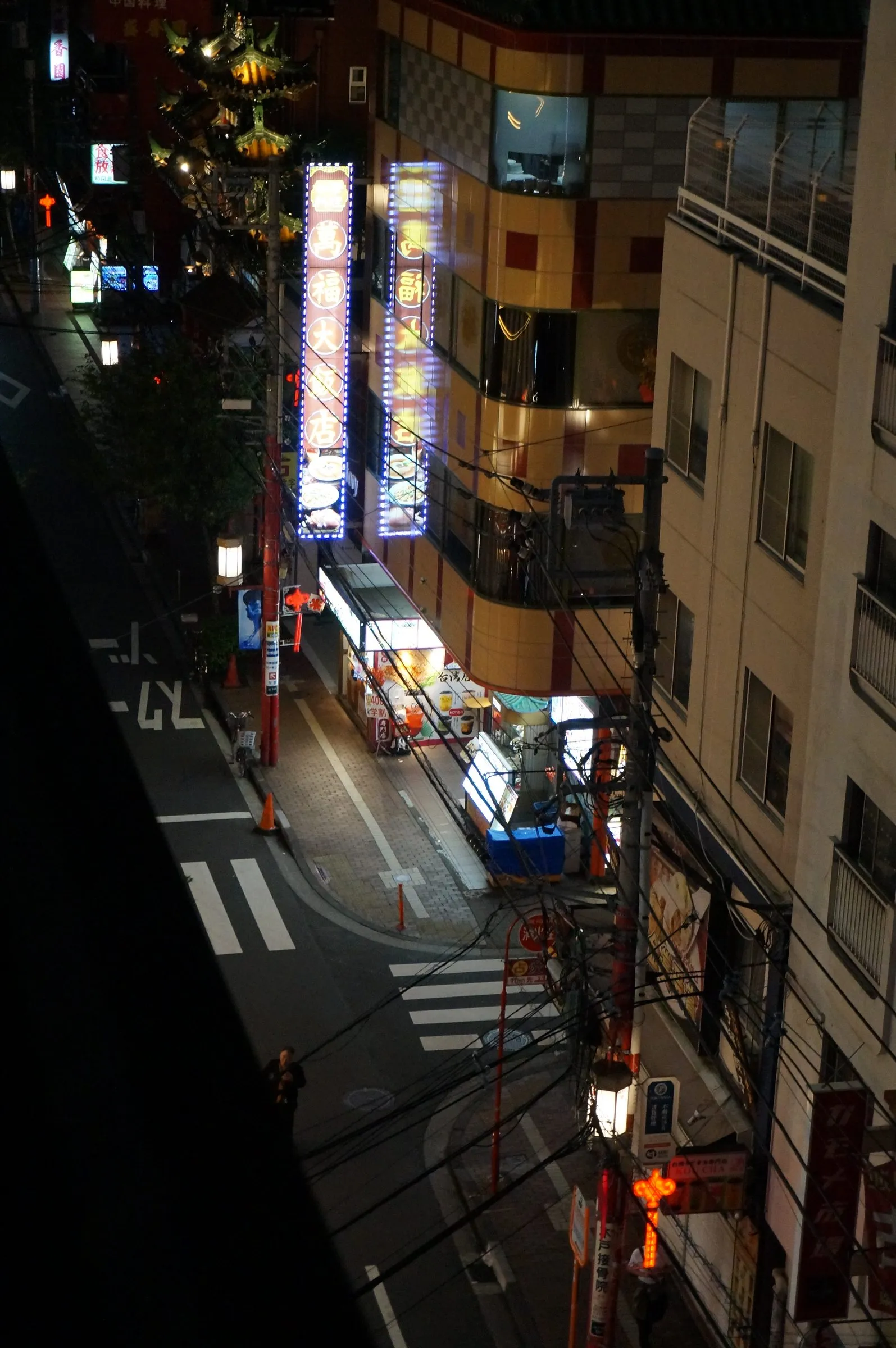 A quiet street at night seen from several floors up, with tall lit signboards, an ornate Chinese-style gate roof and a lone pedestrian at a crosswalk