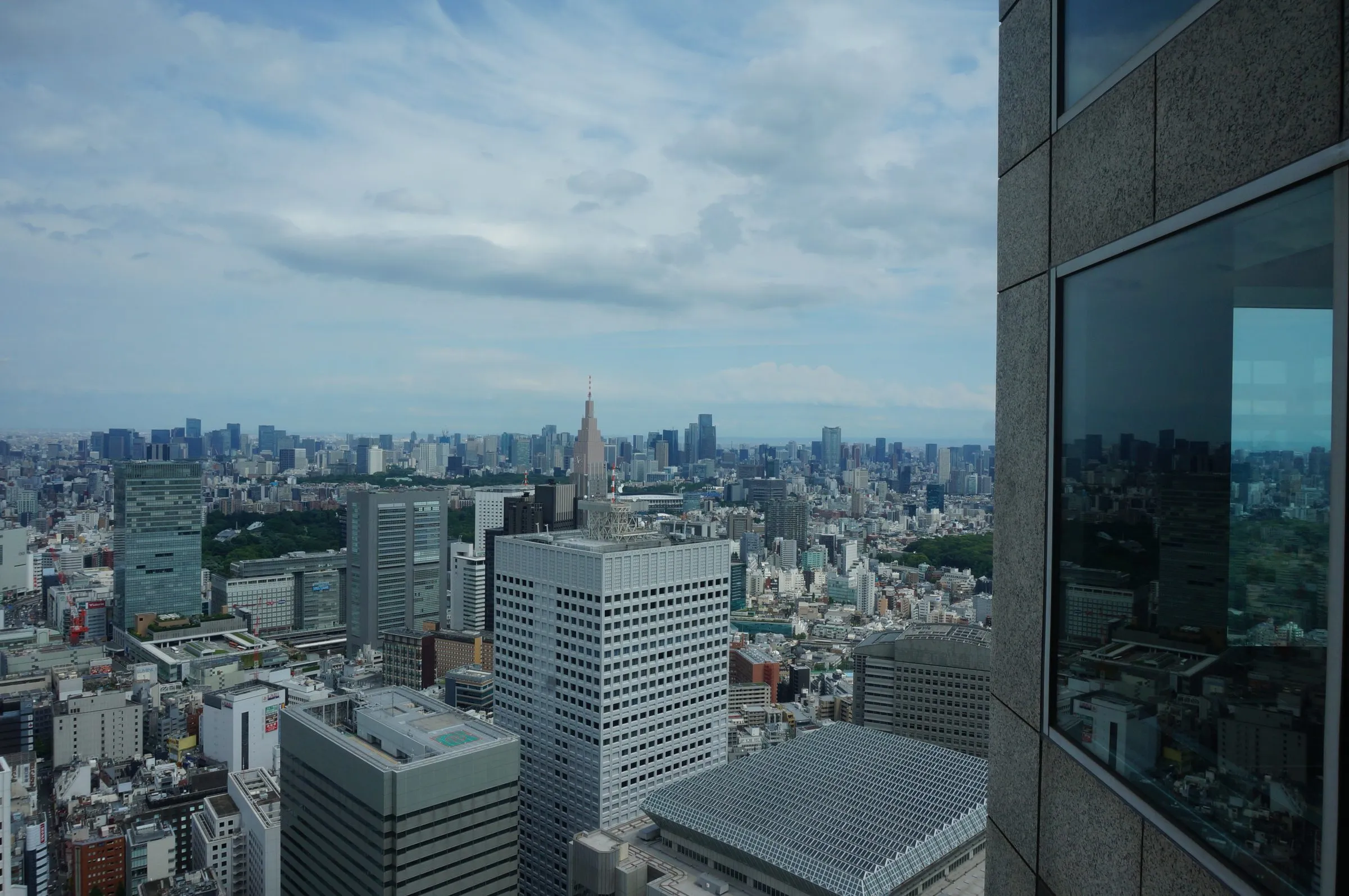 A wide view over Tokyo's office towers and the spired Docomo building to a hazy horizon, the city reflected in a dark window at right