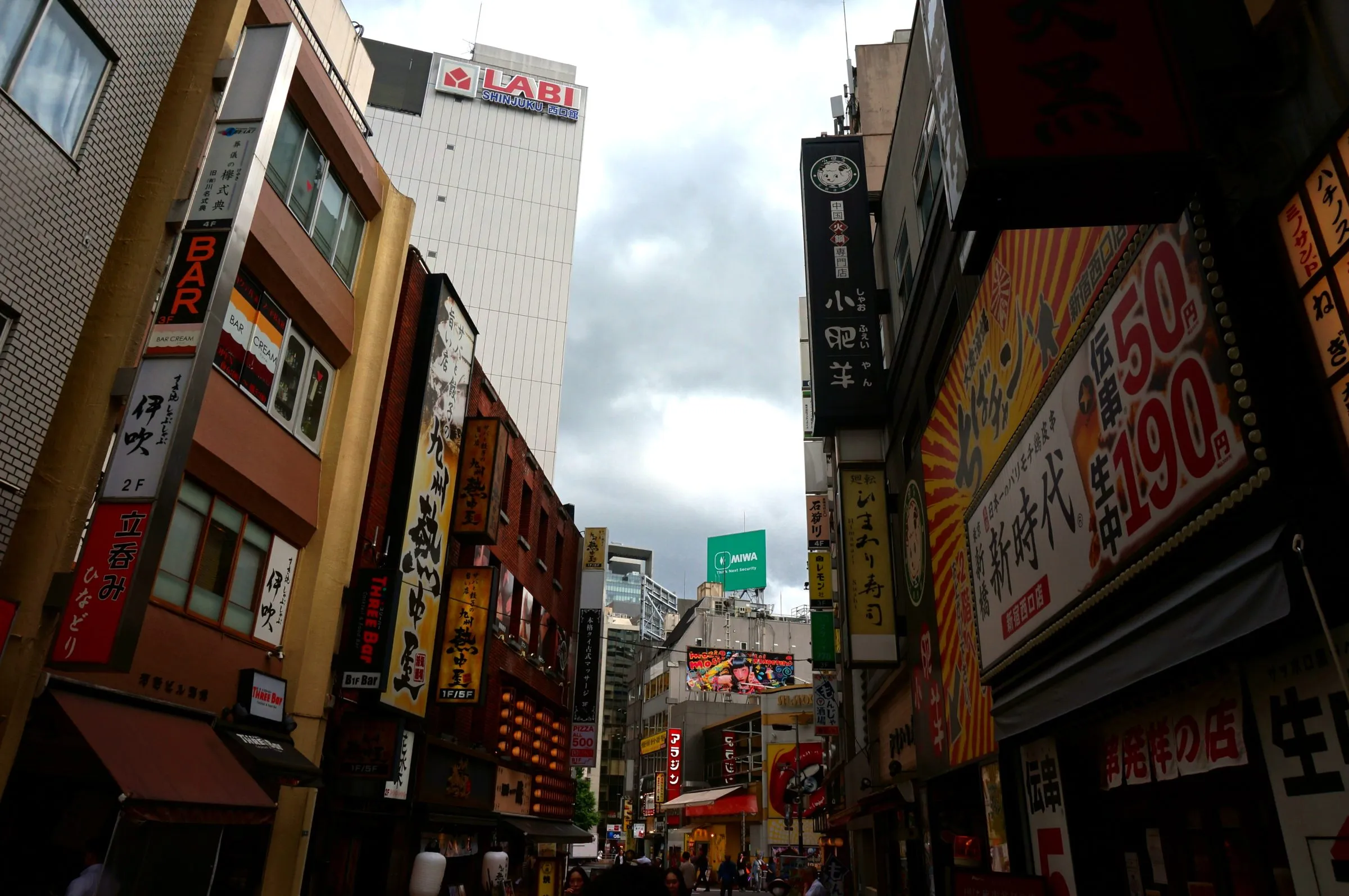A narrow street lined with dense shop signs and neon, seen from below against a cloudy sky