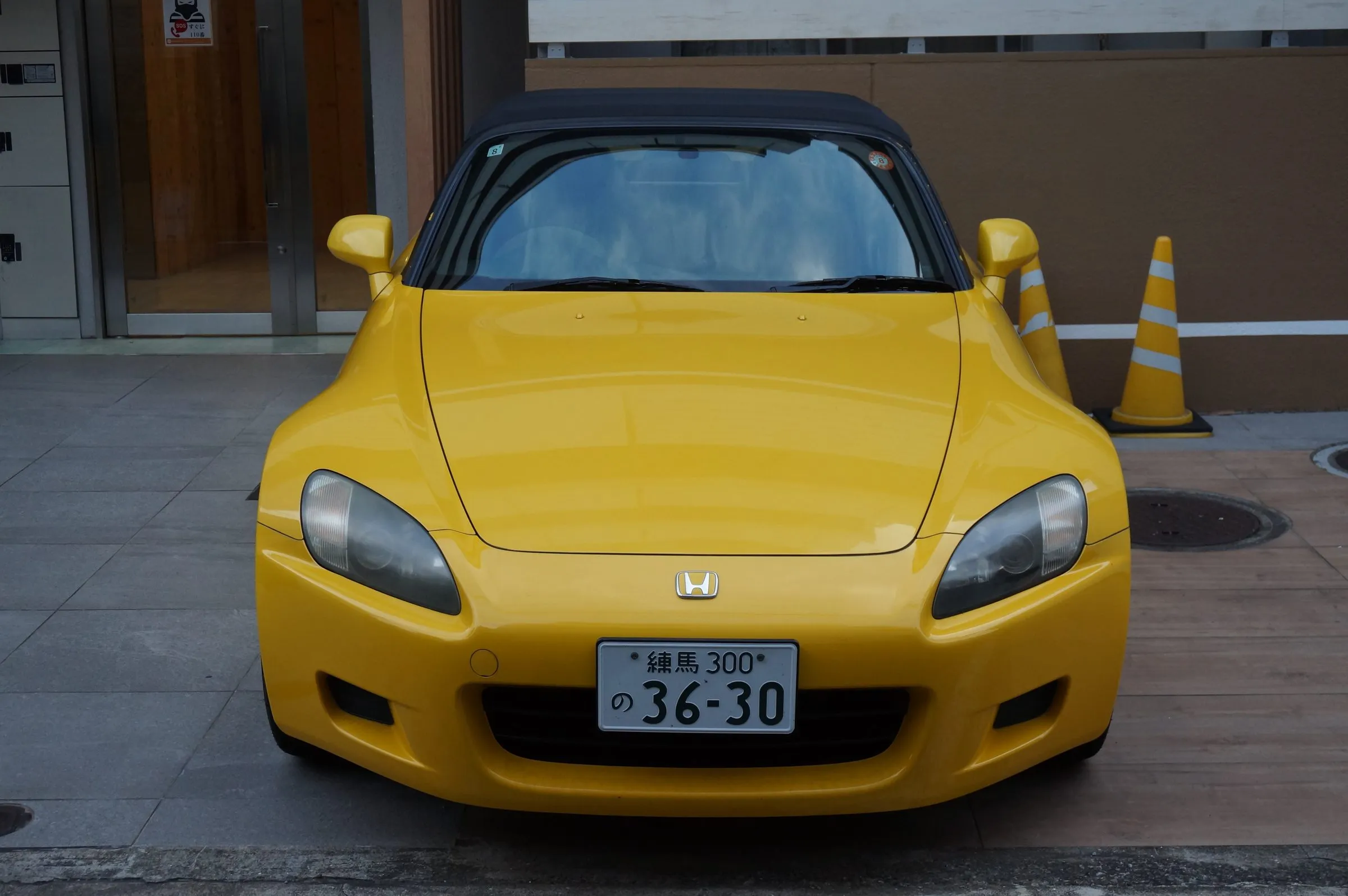 A yellow Honda S2000 roadster with its black soft top up, parked head-on in front of a building with orange traffic cones beside it