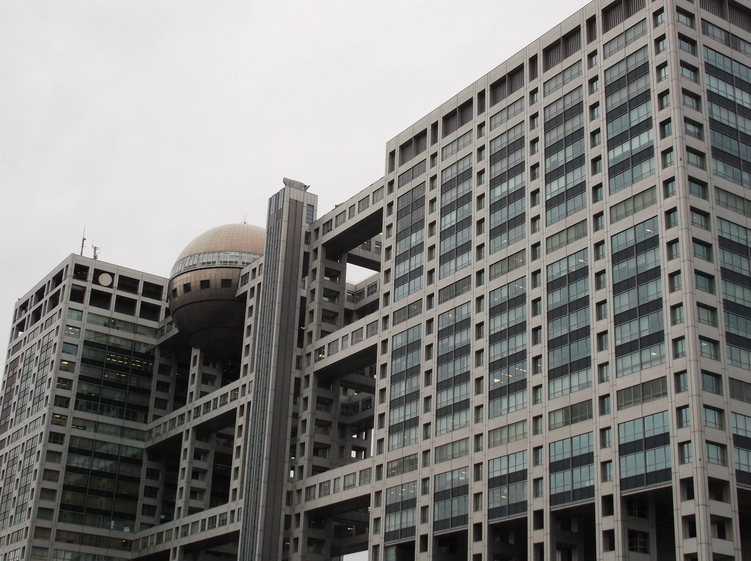 The grid-like Fuji TV building with its bronze sphere suspended between towers under a flat white sky