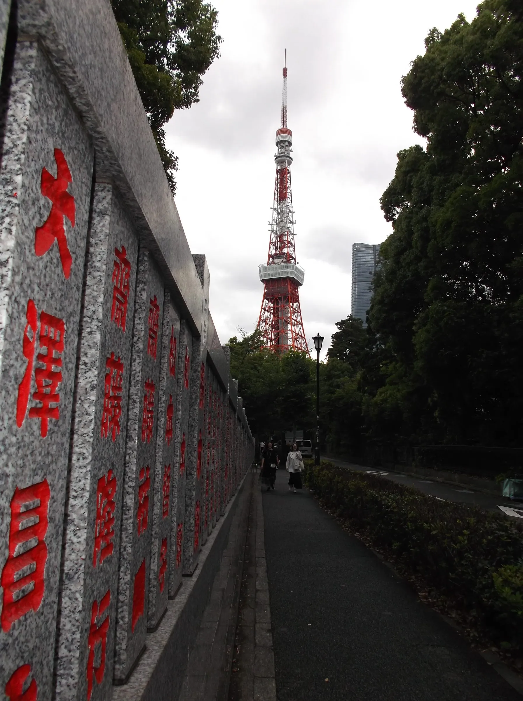 A footpath beside a long granite wall carved with red characters, leading toward the red and white Tokyo Tower behind the trees