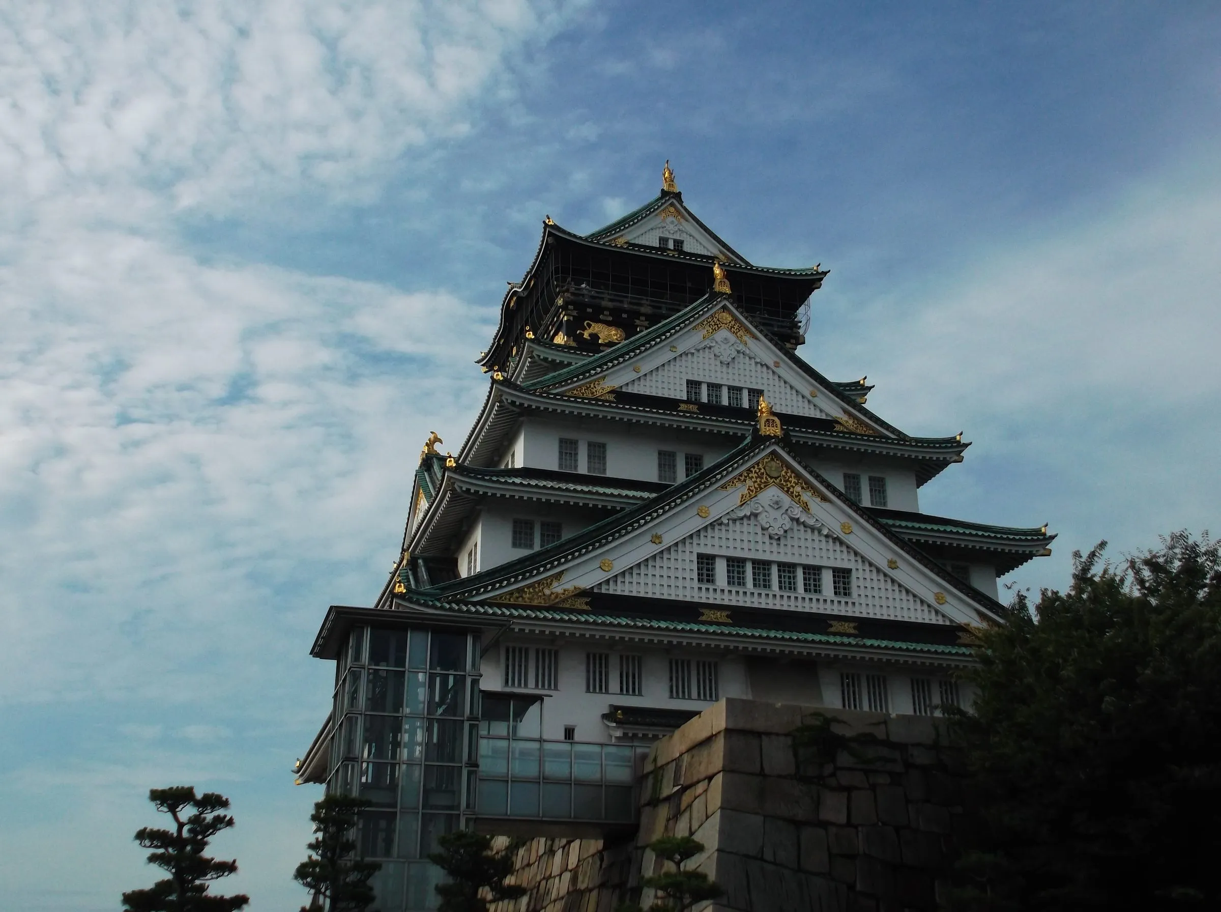 The white and green tiered keep of Osaka Castle with gold ornaments, rising above a stone wall and pines into a blue sky with thin cloud
