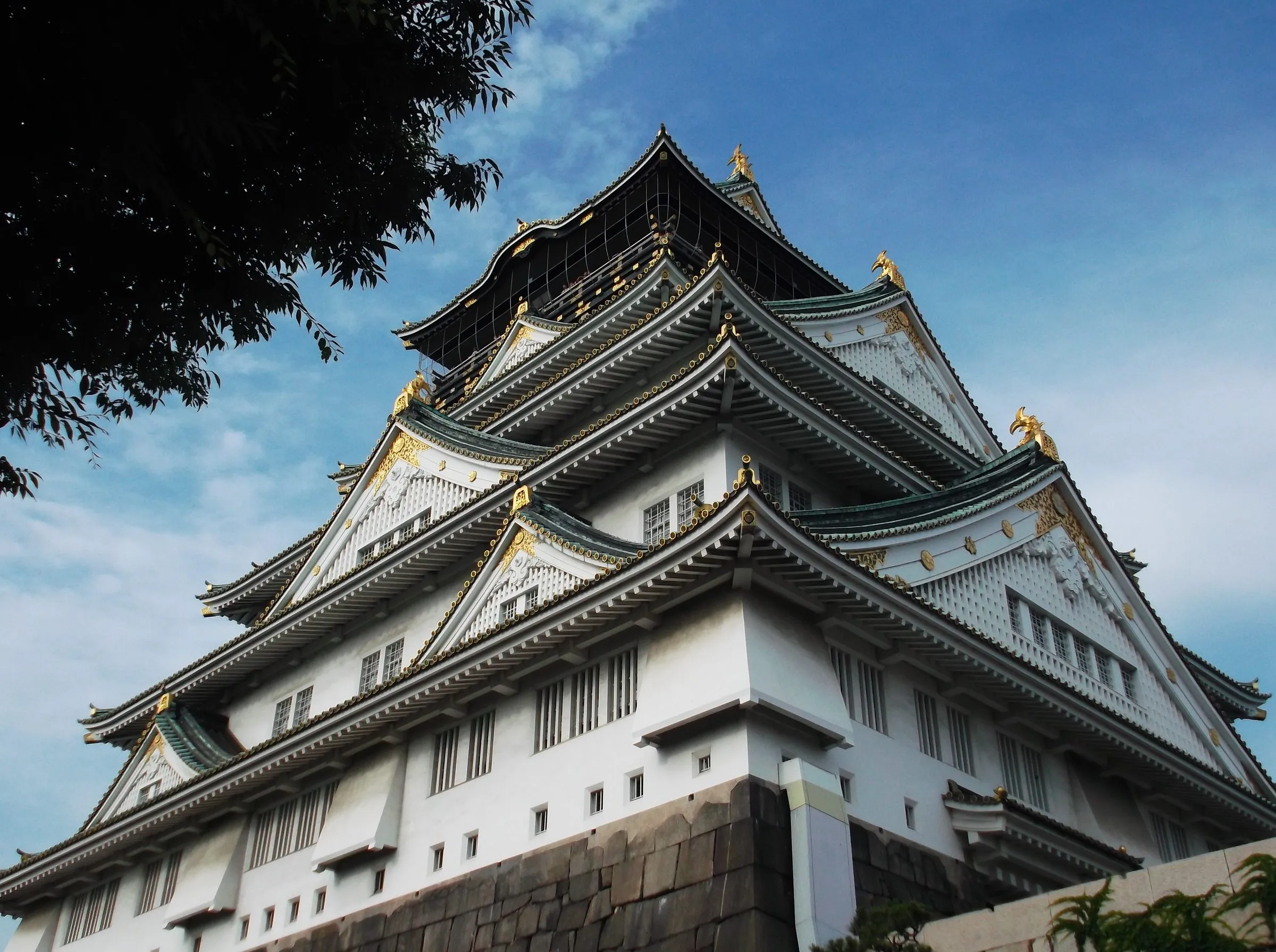 A low-angle view up the layered roofs and gabled walls of Osaka Castle, gilded tigers and fish at the corners, a tree branch overhead