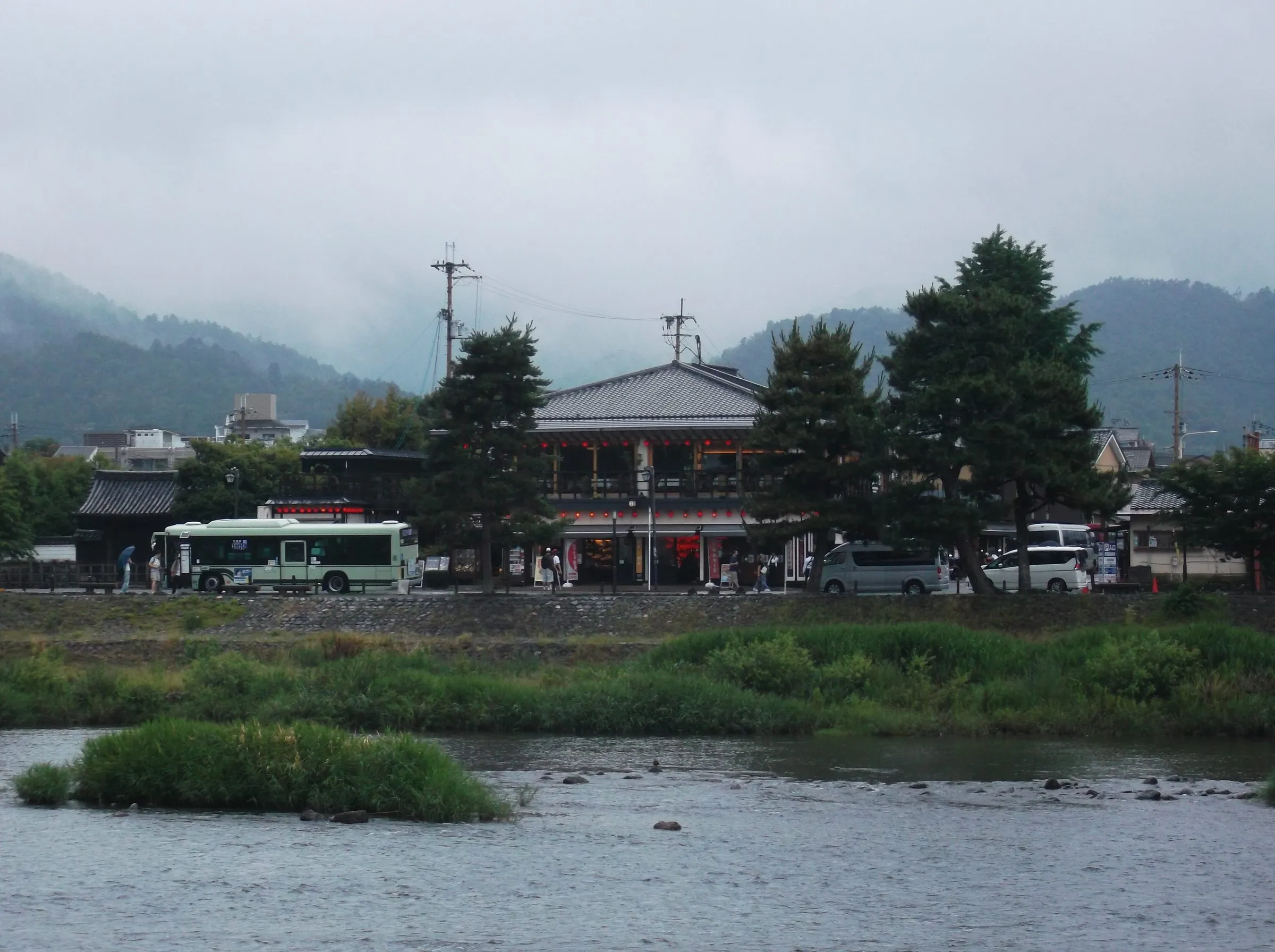 A two-storey shop and a green city bus on the far bank of a shallow river, low mist hanging on the hills behind