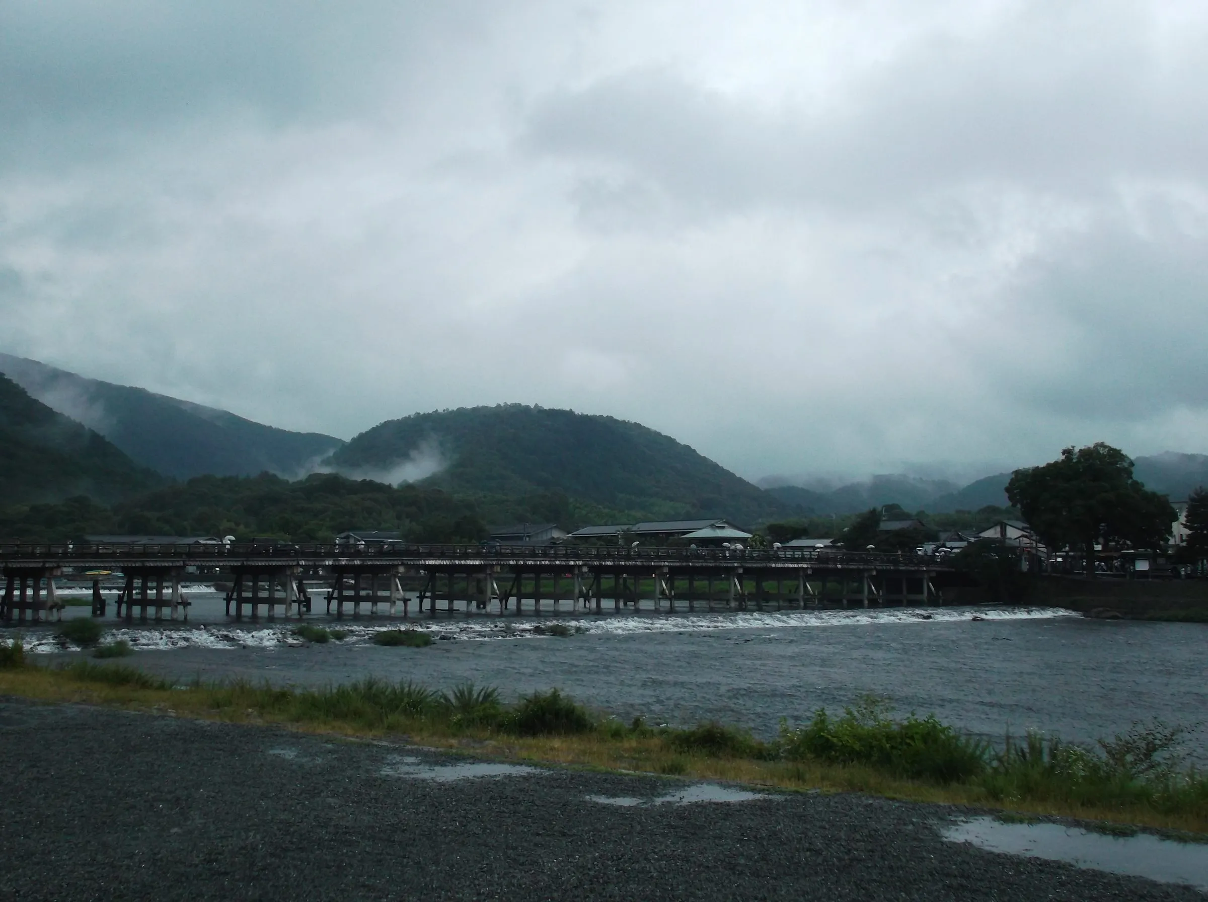 A long wooden bridge across a wide river below a low weir, forested hills wrapped in mist and heavy grey cloud