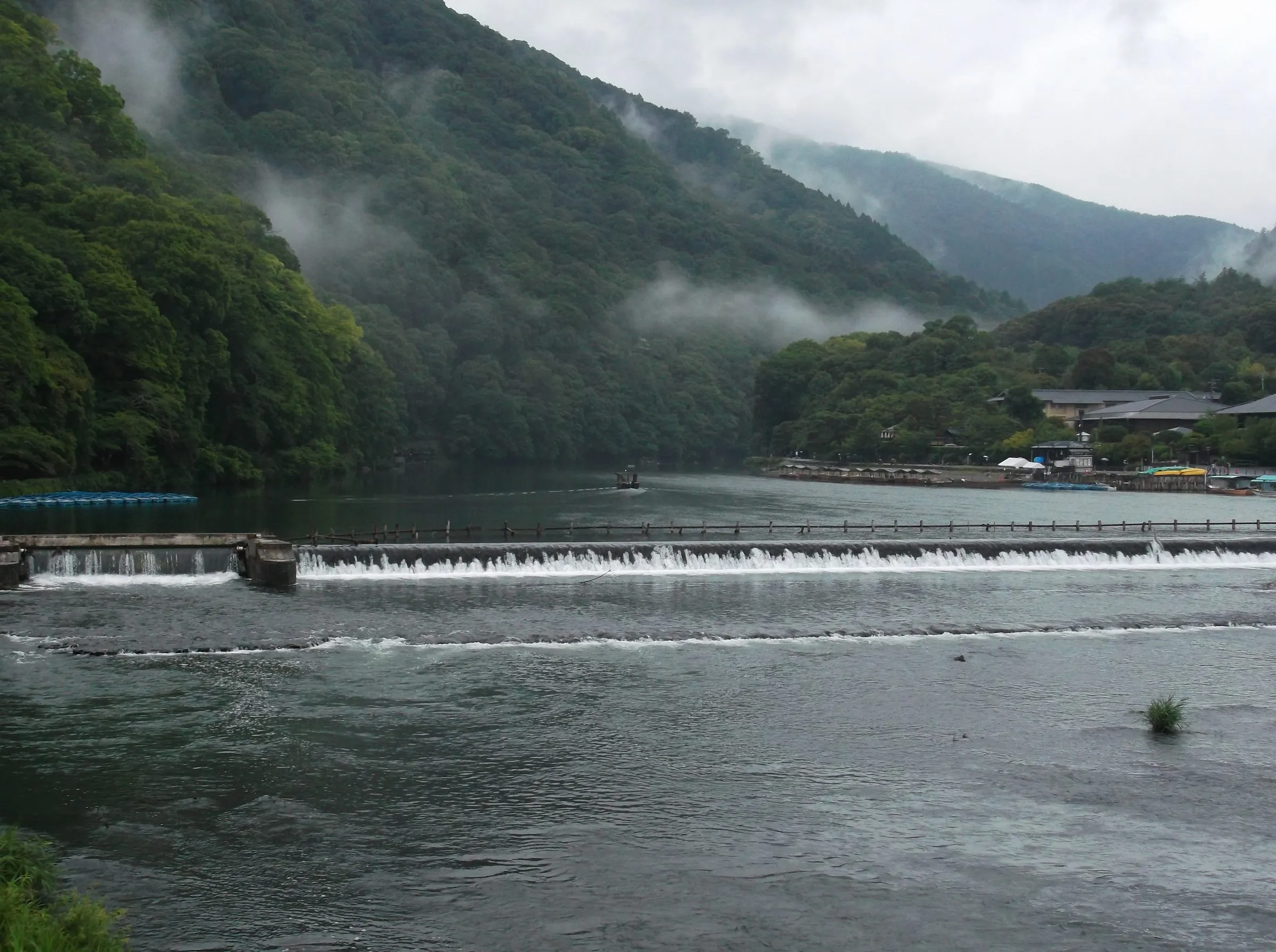 Water spilling over a low weir in a wide river, steep green hillsides with drifting mist rising on either side