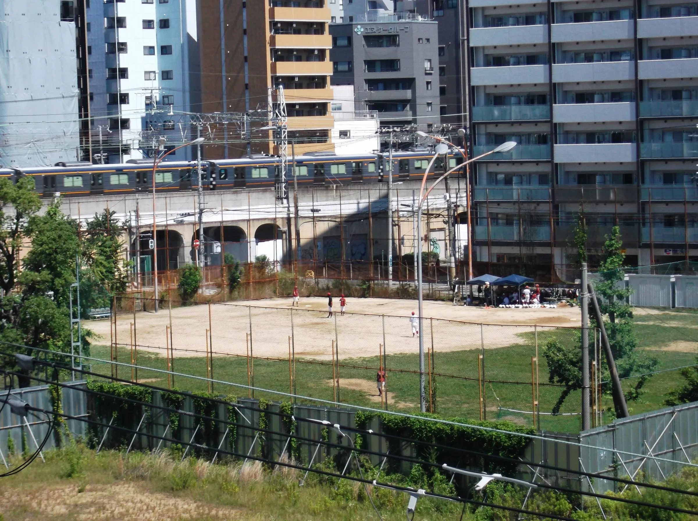A youth baseball practice on a dirt field beside an elevated railway, a passing train and apartment blocks behind
