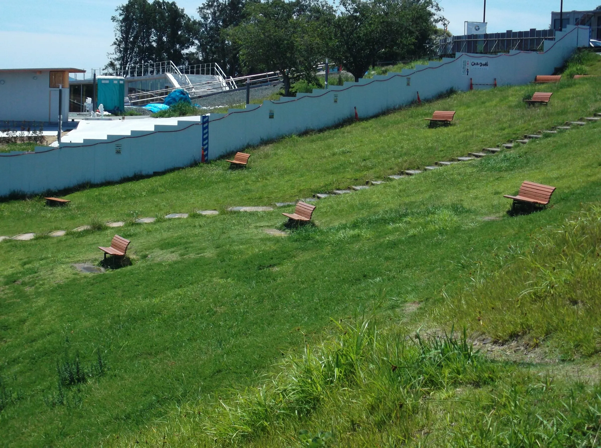 Wooden benches scattered across a grassy embankment, a stepping-stone path climbing toward a white wall and trees