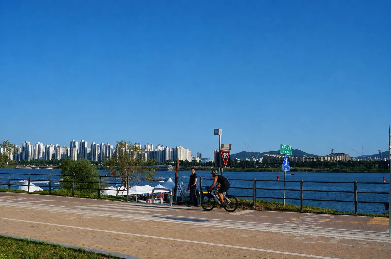 Cyclists on a riverside path along the Han River, apartment towers across the water under a clear blue sky