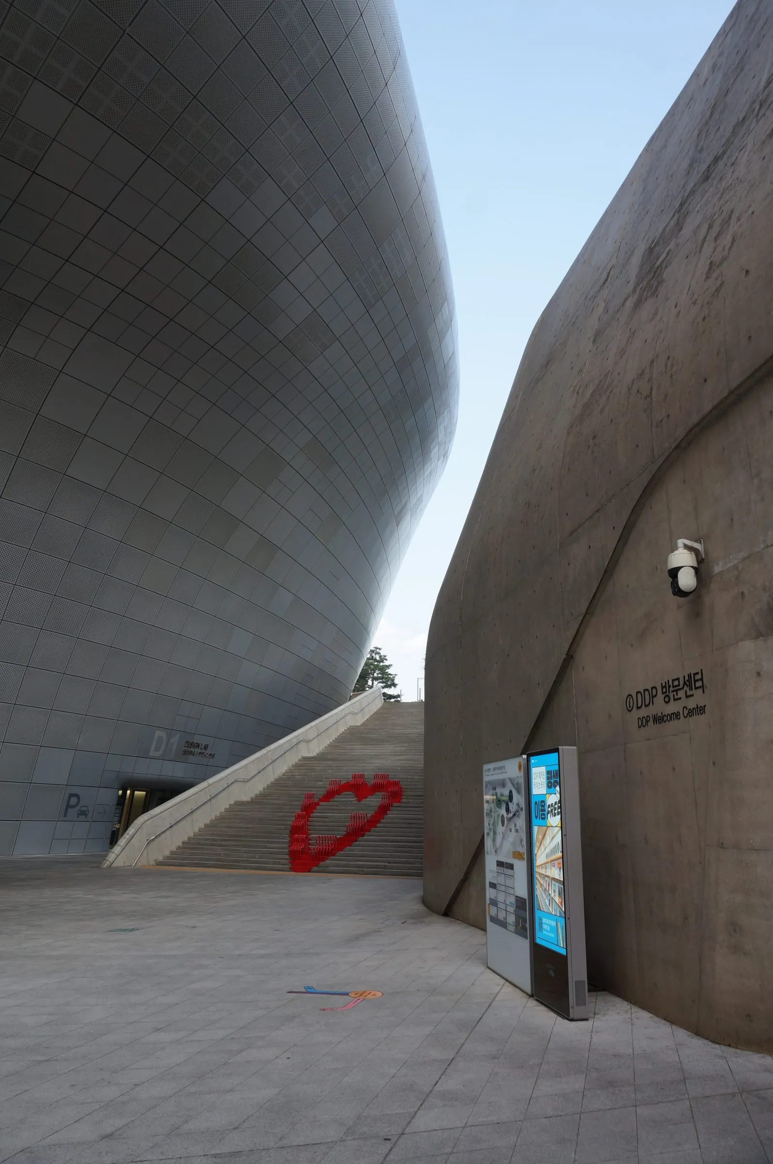 The curving perforated metal facade of Dongdaemun Design Plaza beside a sweeping concrete wall, a red heart sculpture on the steps between them.