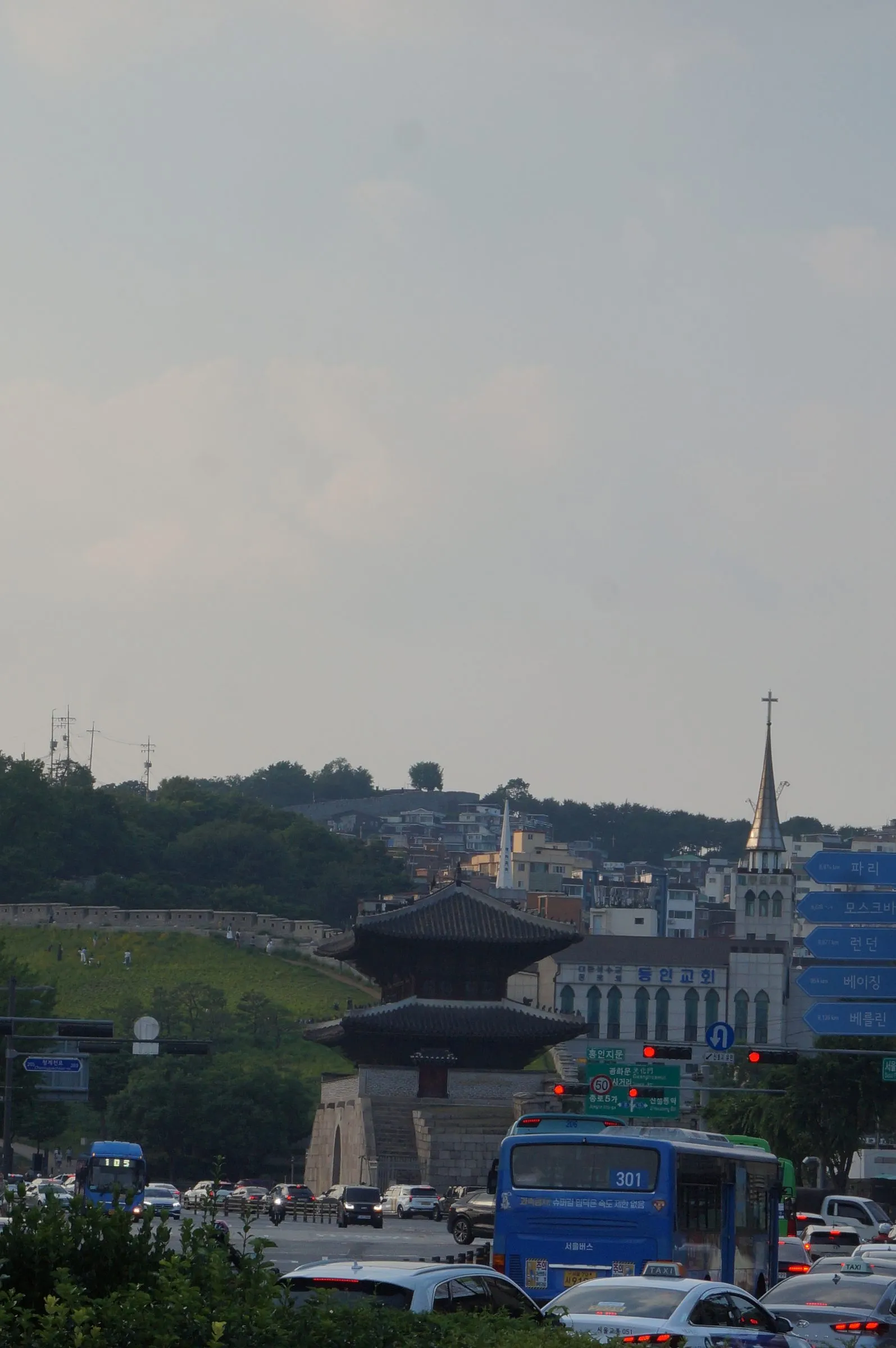 Heunginjimun gate with its two-tiered roof among buses and cars, the old city wall climbing a green hill and a church spire behind.