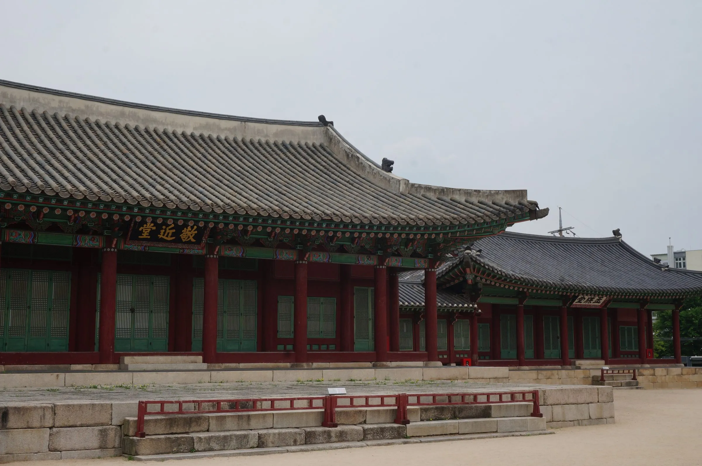 A traditional palace hall with red columns, painted eaves and a curved tiled roof, raised on a stone terrace under an overcast sky.