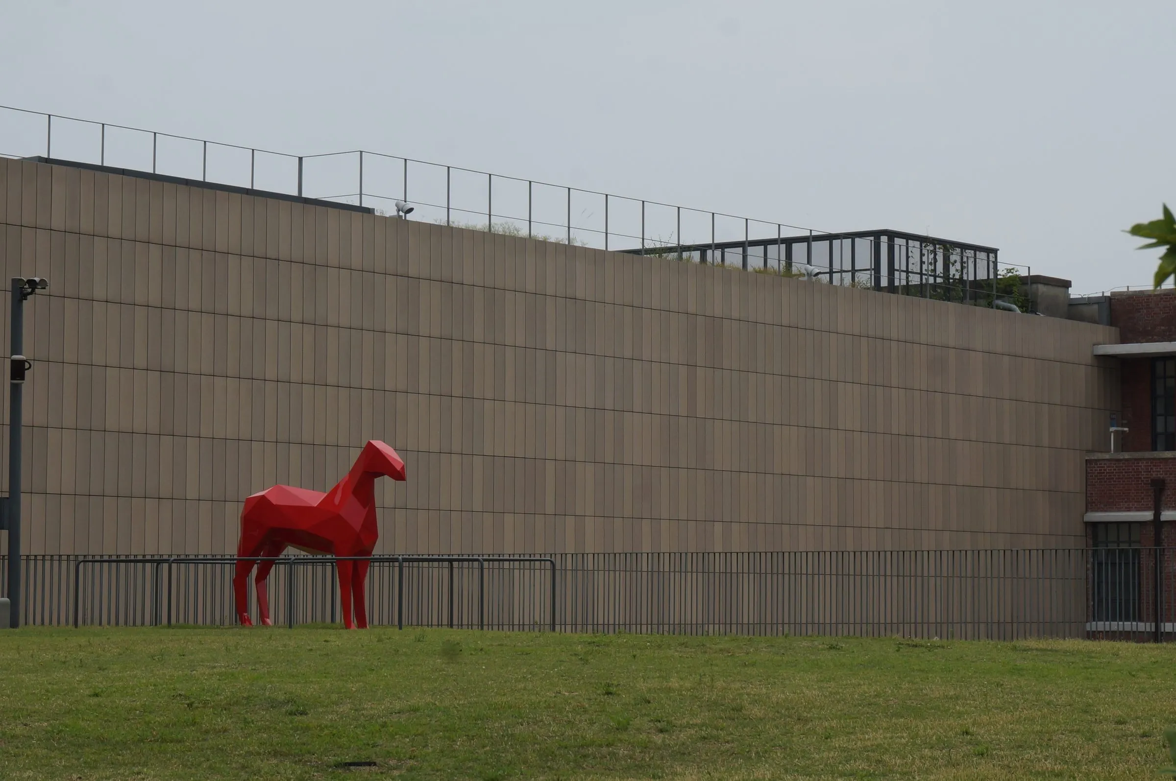 A faceted red horse sculpture standing on a grass lawn in front of a long, plain tan wall.