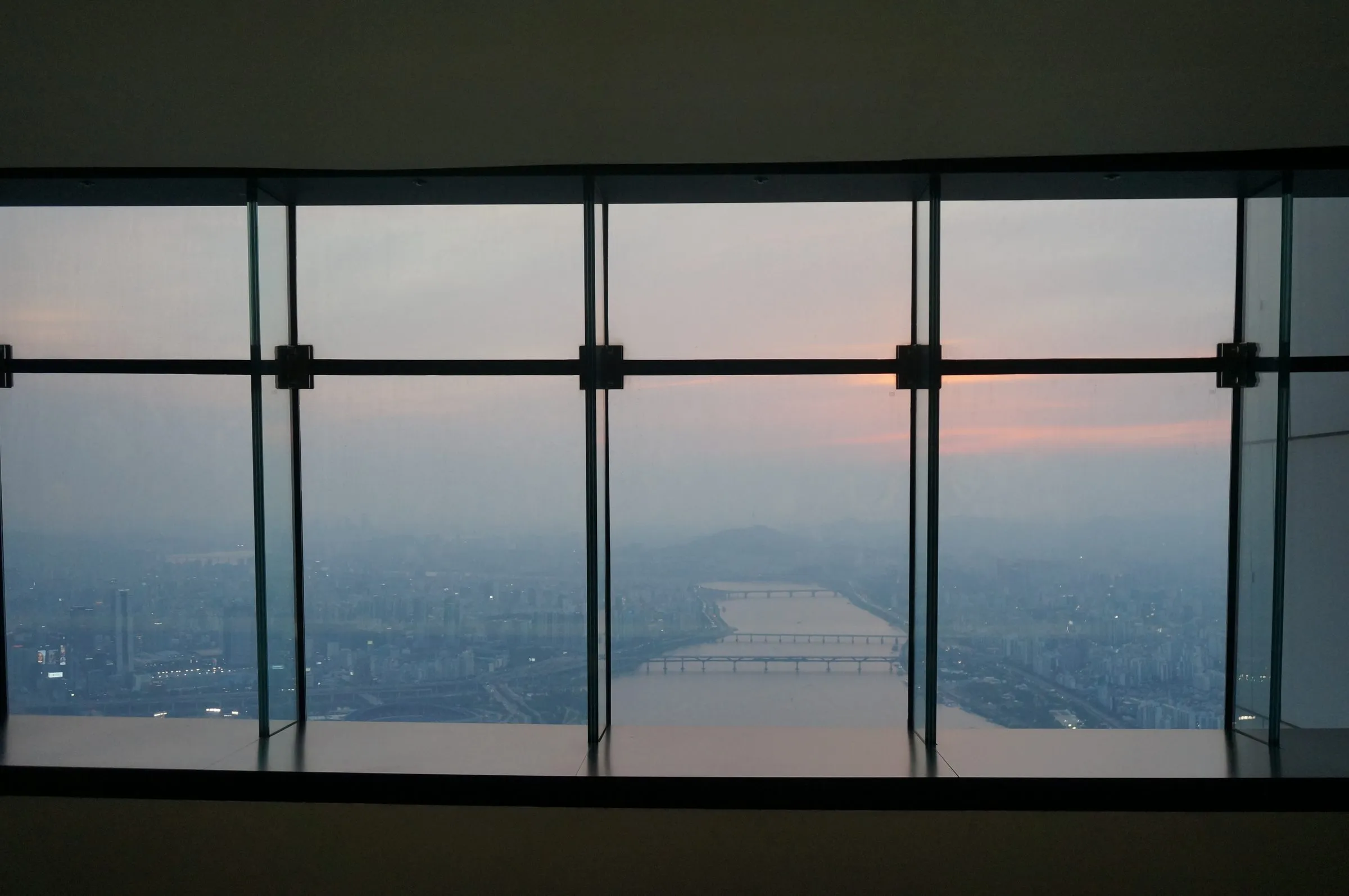 The view through tall observation-deck windows high above Seoul, the Han River and its bridges fading into haze under a pink sunset.