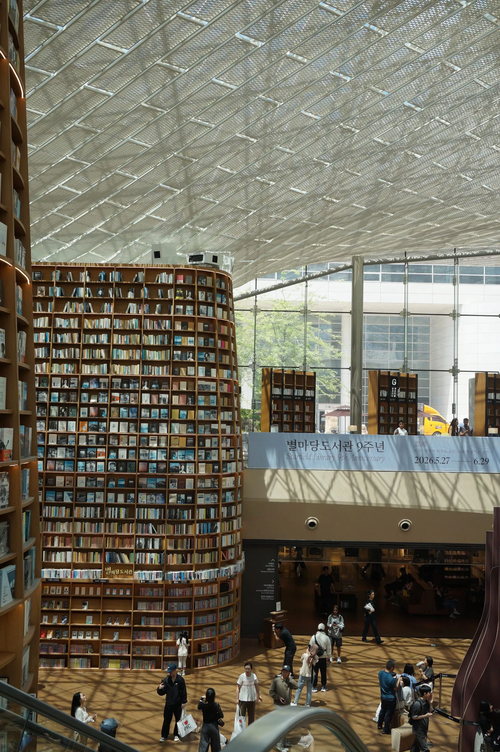 Towering curved bookshelves rise beneath a latticed glass roof at Starfield Library, with people crossing the floor below.