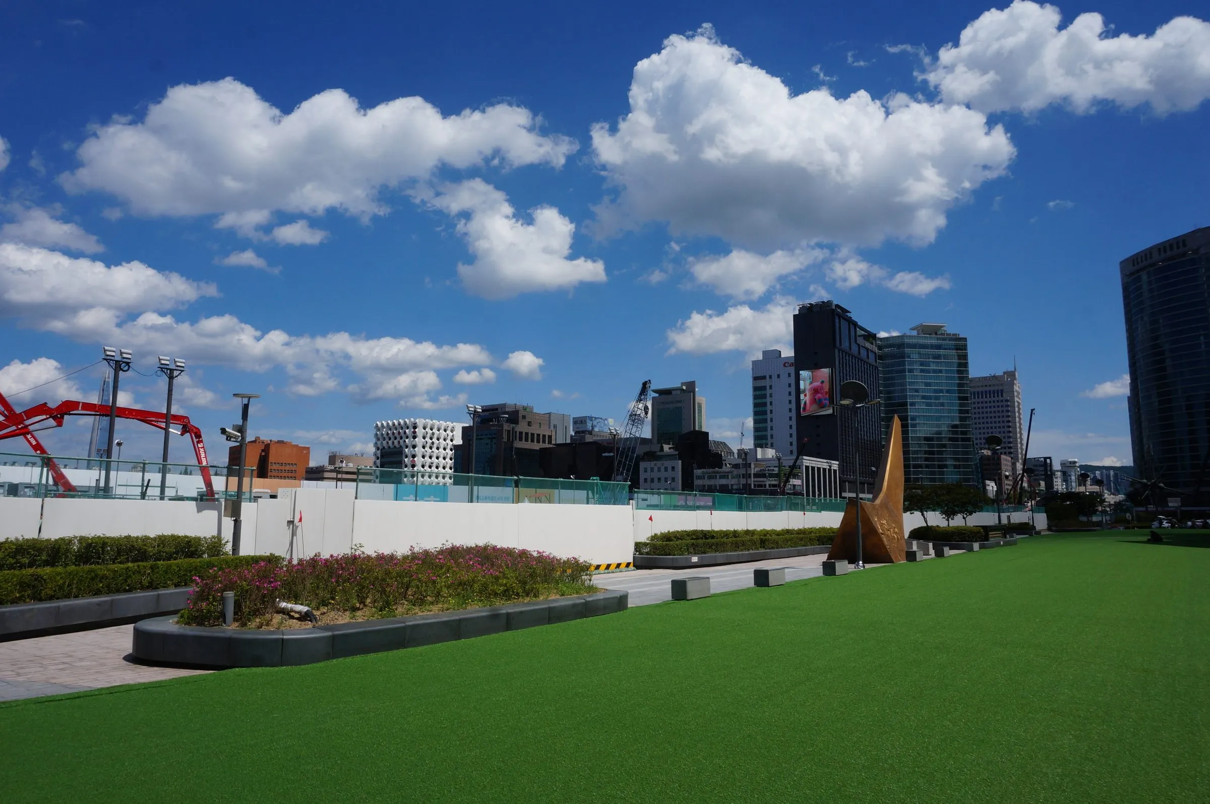 A bright green rooftop lawn with a bronze sculpture, office towers and construction cranes beyond under a blue sky with big white clouds.