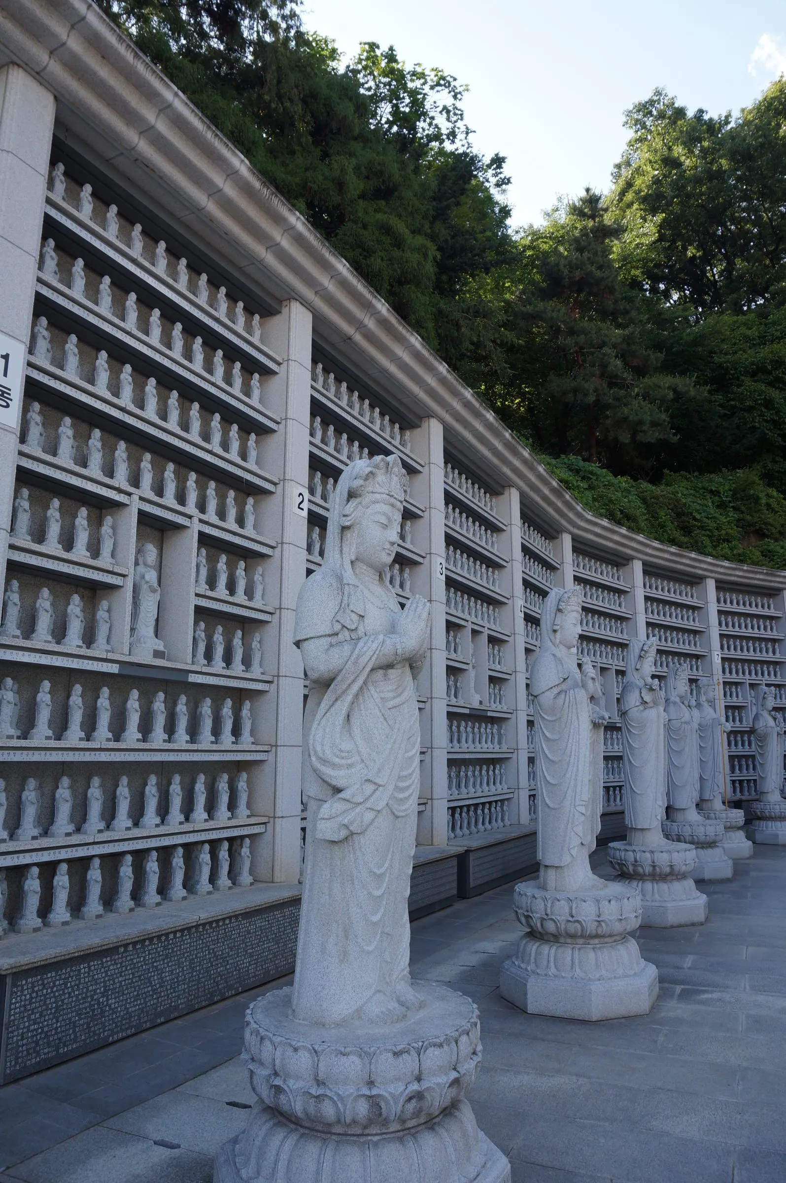 Stone bodhisattva statues on lotus pedestals stand before a curved wall lined with hundreds of small carved figures, trees above.