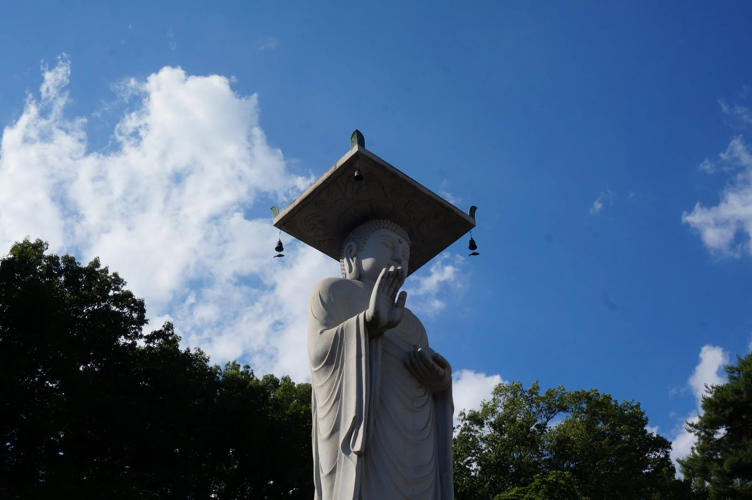 A towering stone Buddha with a square stone canopy hung with small bells, seen from below against blue sky and white cloud.