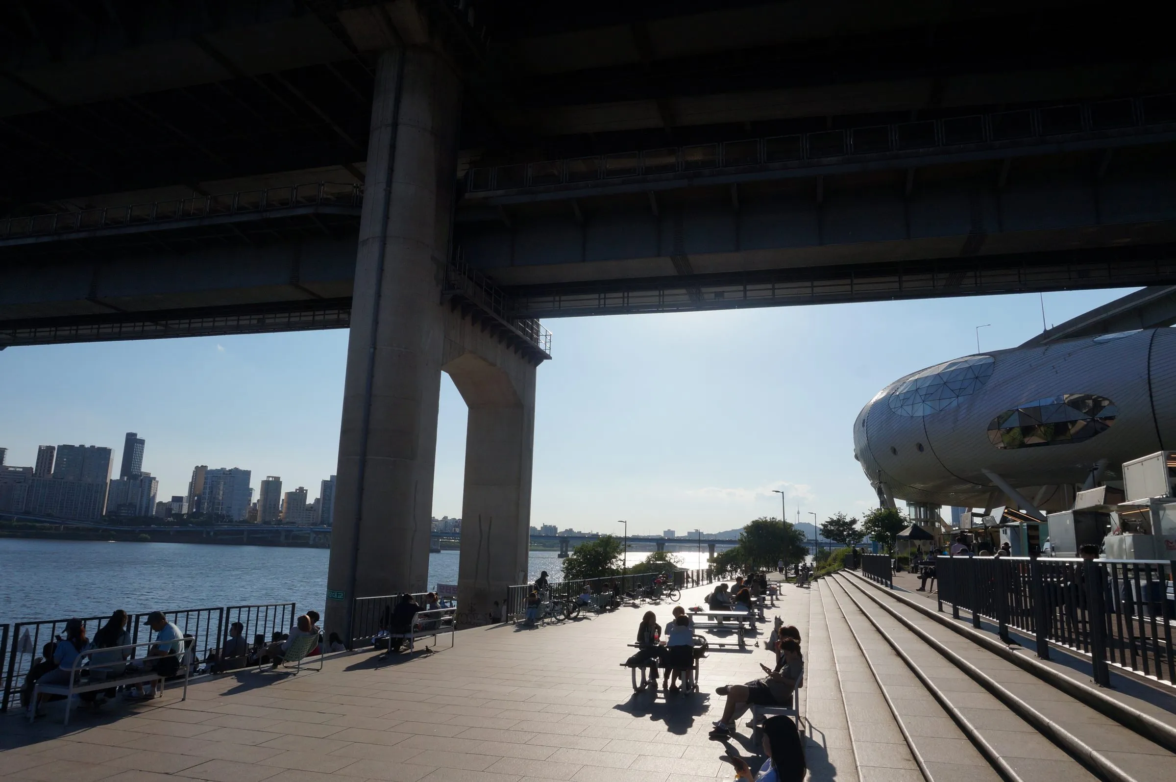 People sit in the shade beneath a double-deck bridge on the Han River, a silver tube-shaped building to one side and apartment towers across the water.