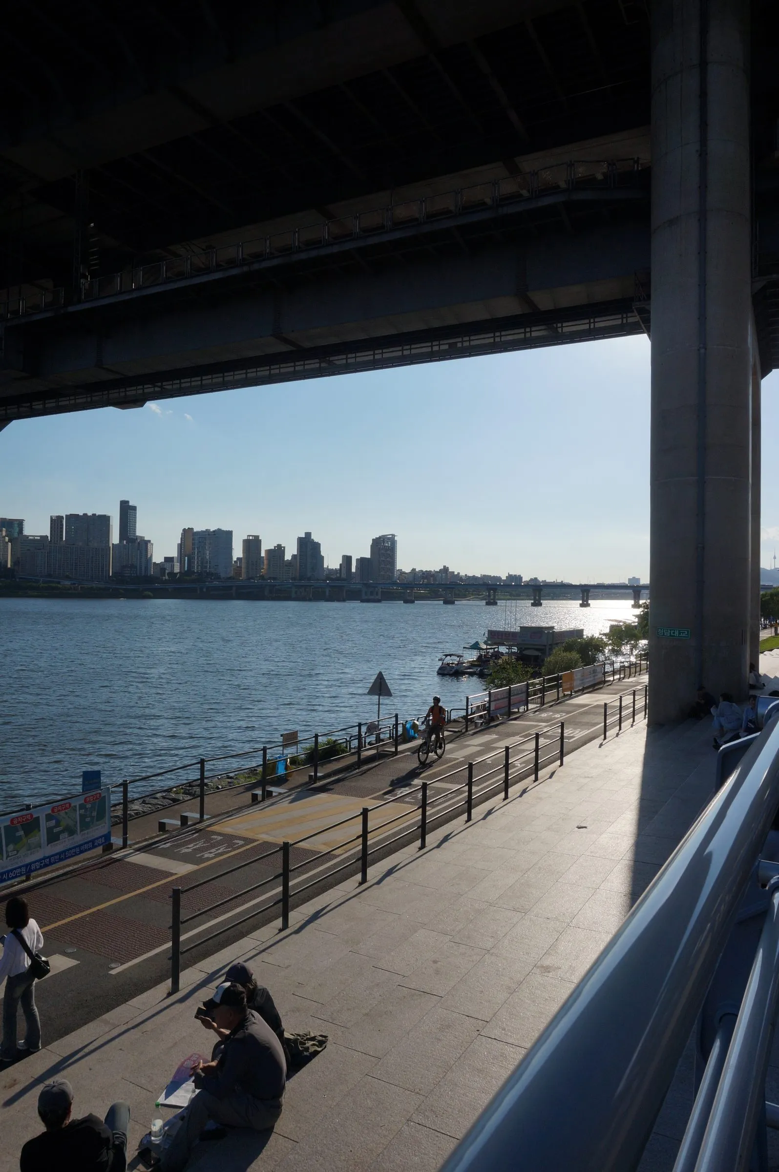 A cyclist rides the riverside path below a bridge deck while people sit on the steps in the foreground, the Han River and far-bank towers in the sun.