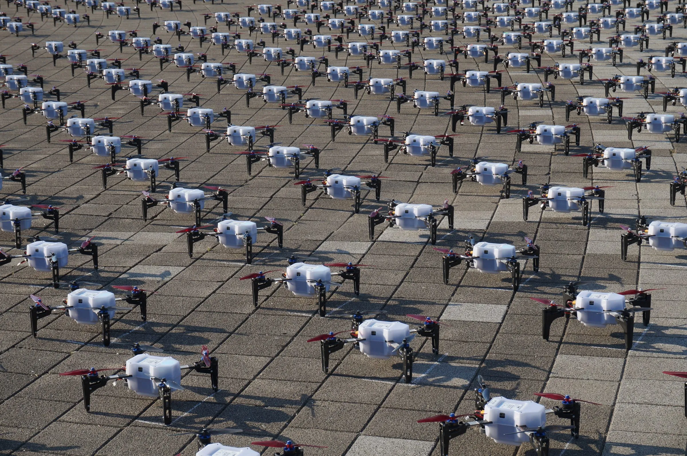 Hundreds of white drones with red propellers laid out in a grid on paving stones.