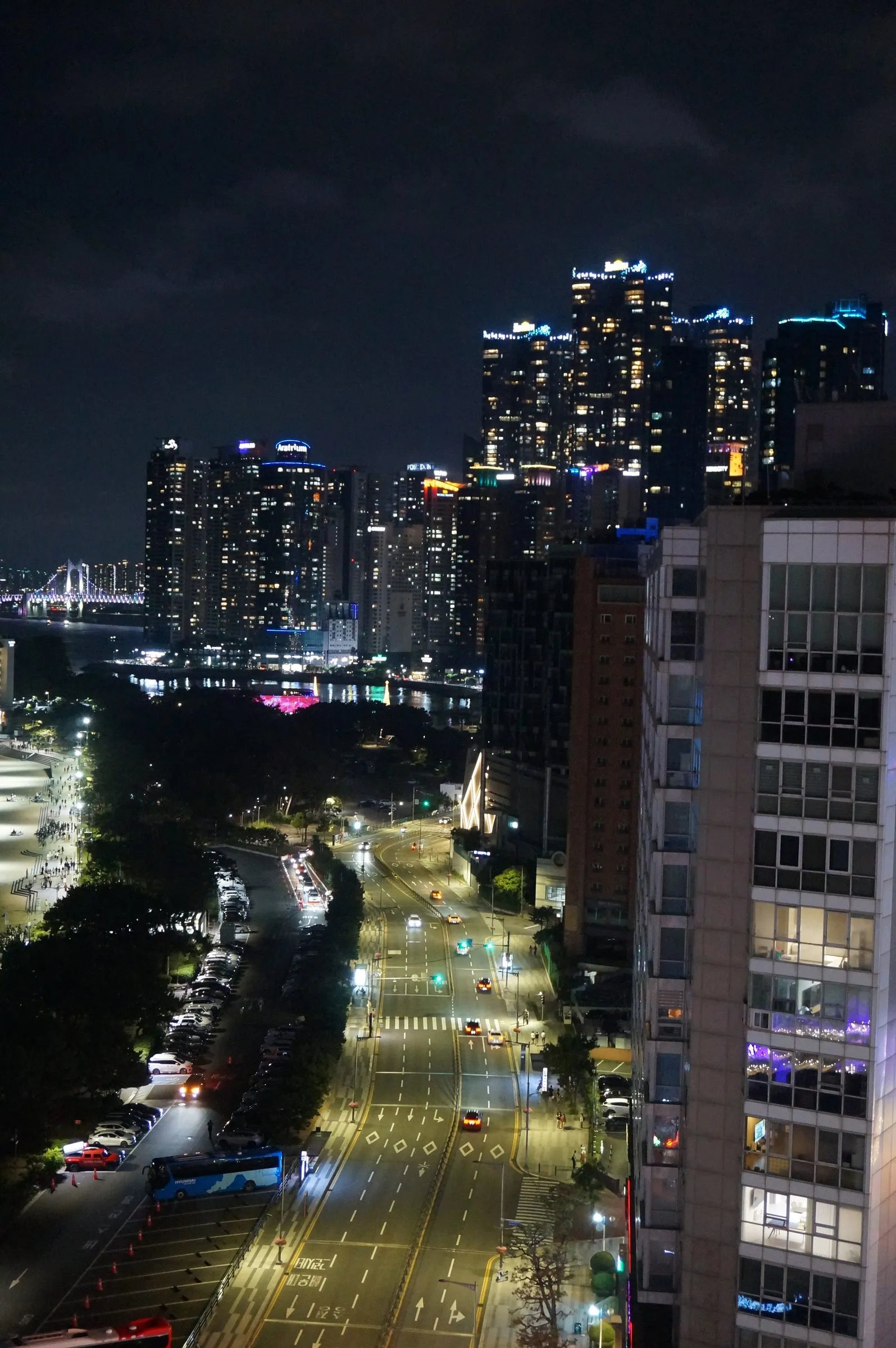 A night view down a lit avenue toward clustered high-rise towers by the water, a glowing suspension bridge on the far left.