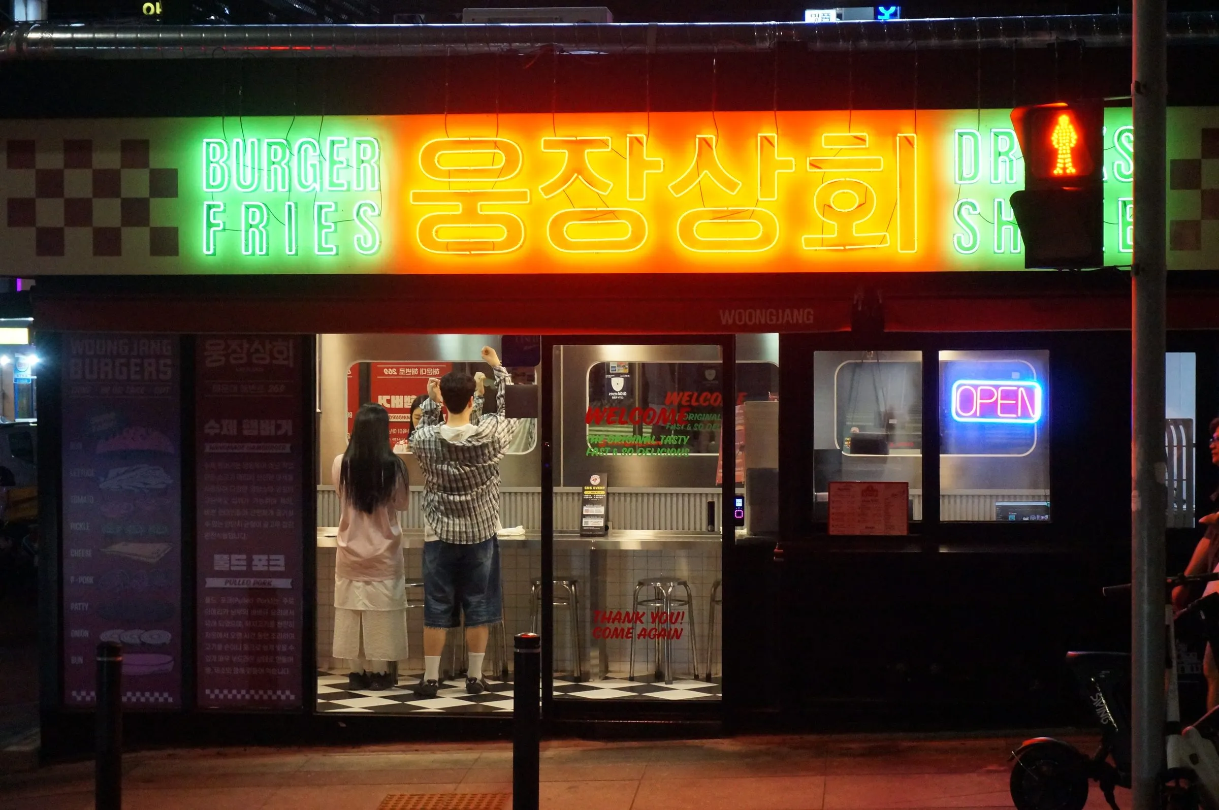 Two people at the counter of a burger shop under a glowing green and orange neon sign in Korean, a red pedestrian signal lit beside it.