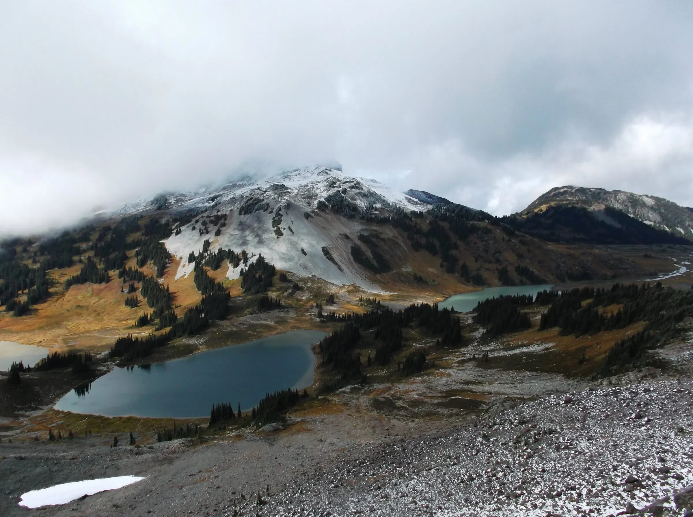 A snow-covered peak half hidden in cloud above a basin of alpine lakes and autumn meadow, frosted scree in the foreground