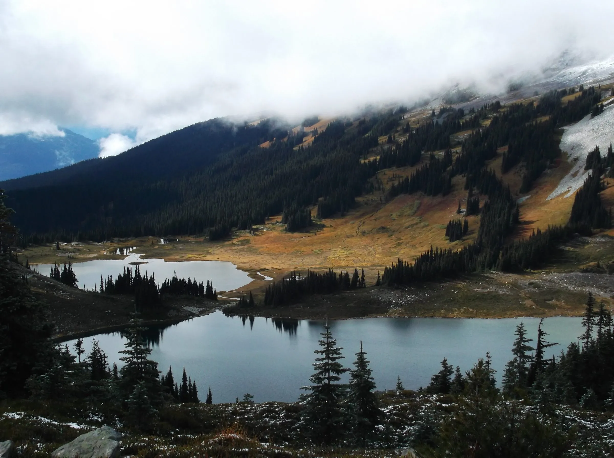 Two alpine lakes in a forested basin below a mountainside, low cloud resting on the ridge