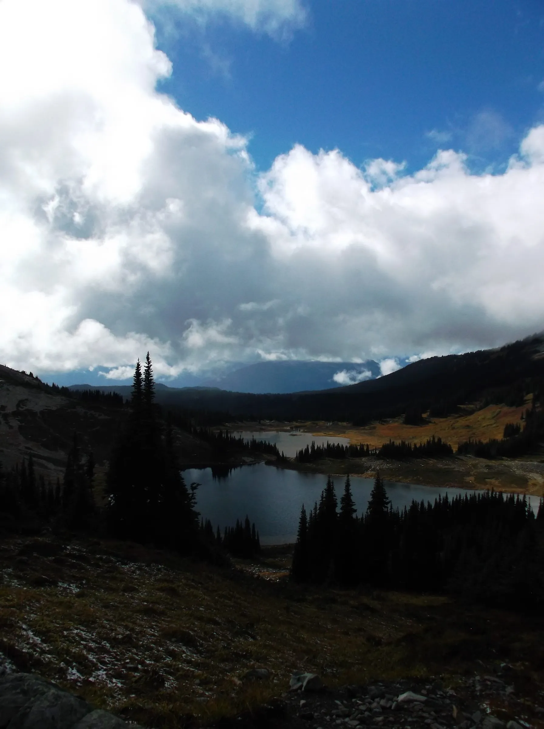 Billowing white clouds parting to blue sky above two lakes in a forested valley, silhouetted firs in front