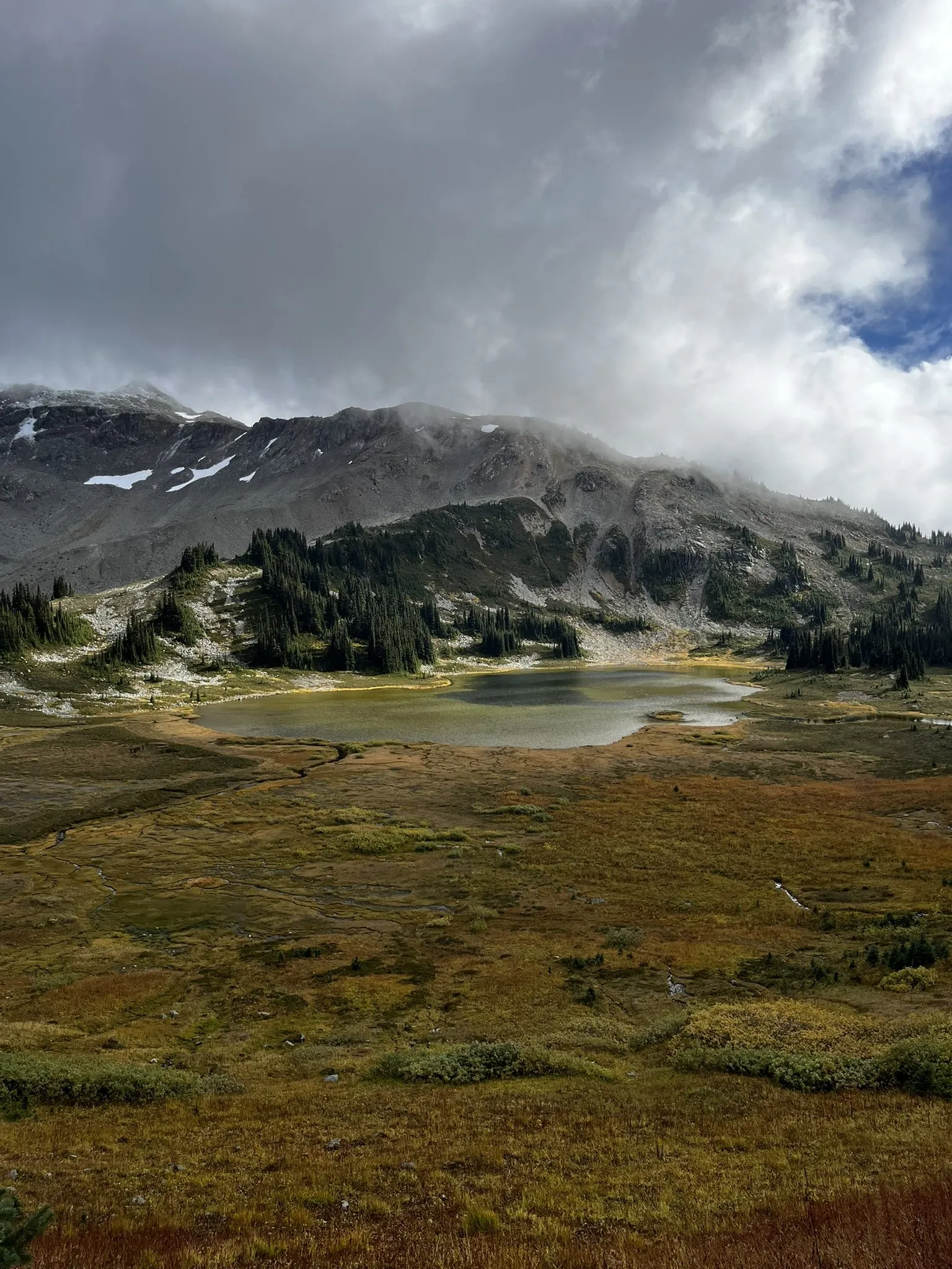 An autumn alpine meadow with a small lake, snow-dusted peaks and heavy clouds above
