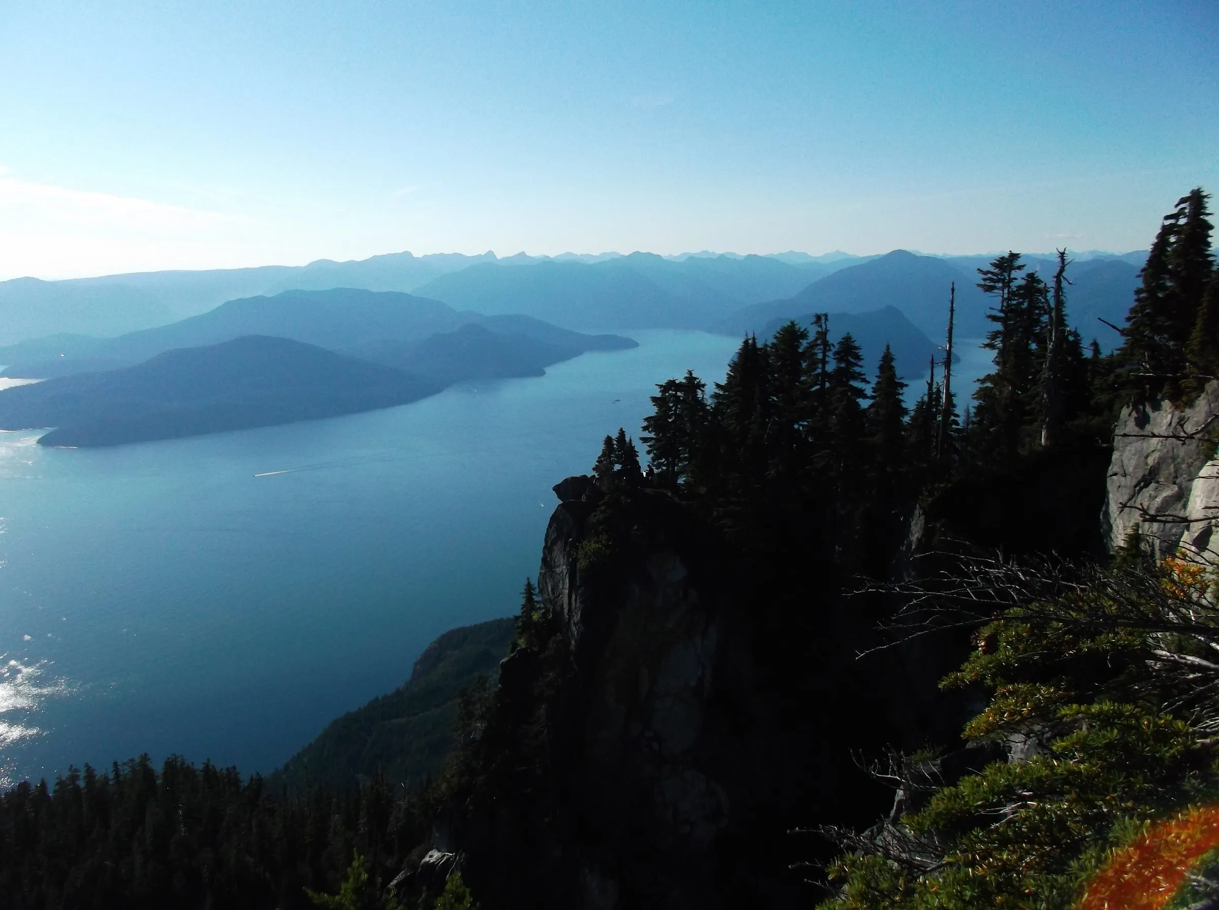 A forested rock bluff dropping toward the blue water of Howe Sound, layered islands and mountains fading into haze