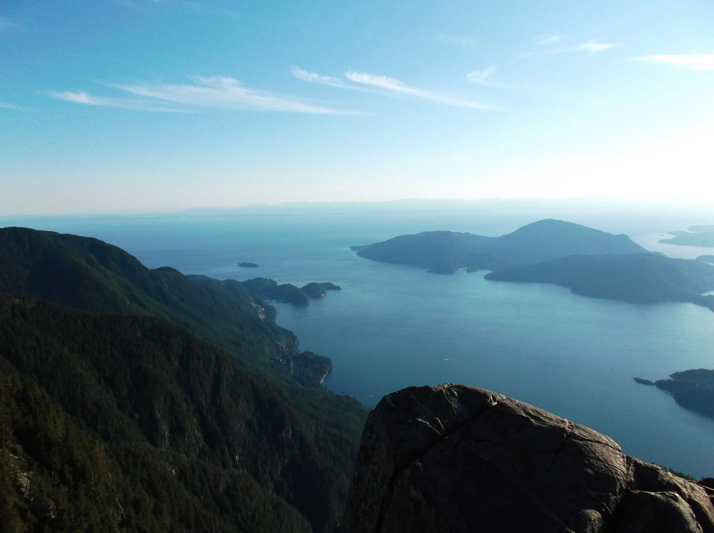 A granite boulder in the foreground above forested slopes, Howe Sound and its islands under thin wispy cloud